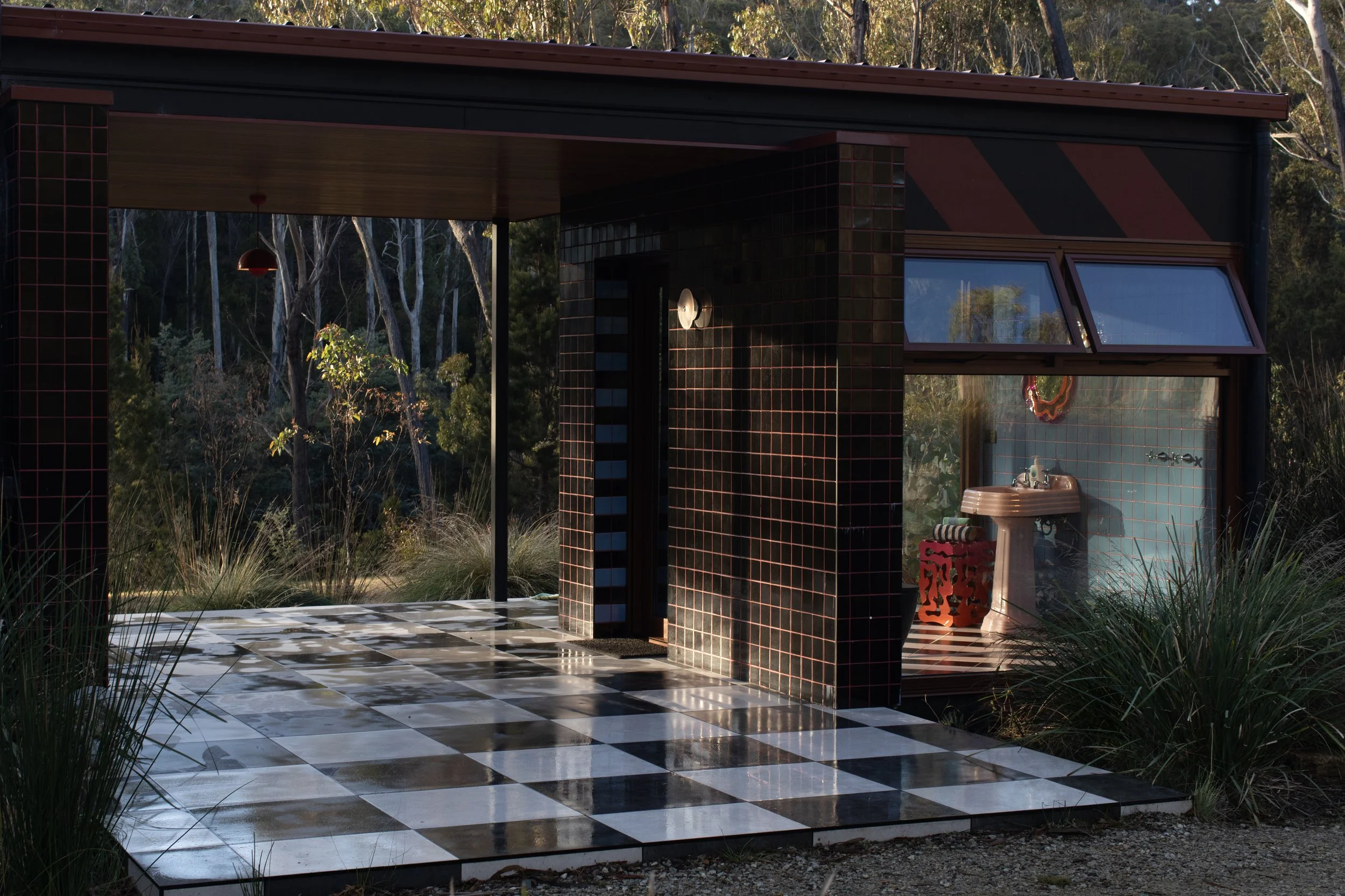 An outdoor shower with black tiled walls and a roof, situated in a natural setting surrounded by trees and tall grass, with a black and white checkerboard-patterned floor.