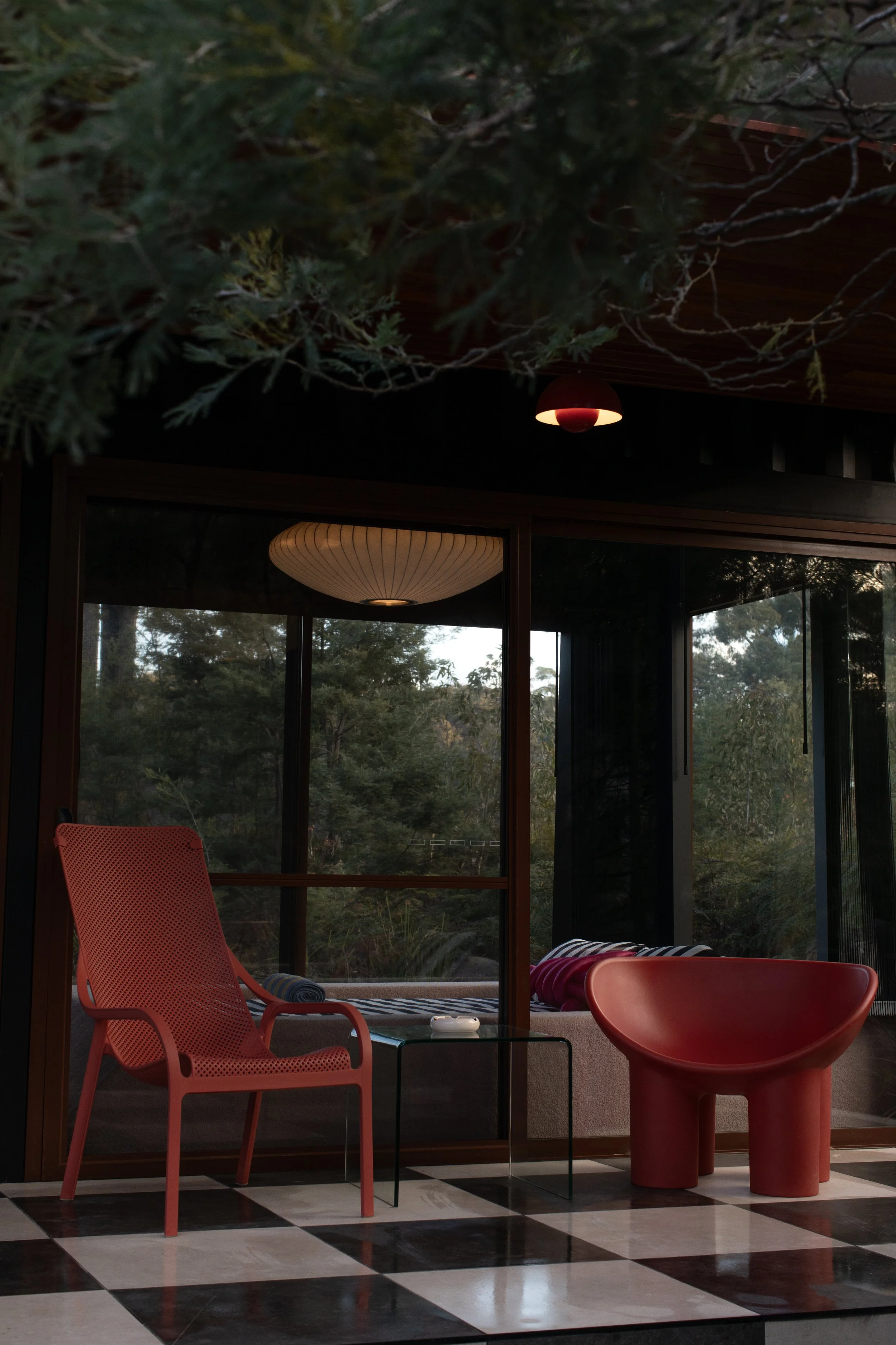 View of indoor seating area with a red chair and large red chair, a small black table, near sliding glass doors with a view of trees and outdoor landscape, and ceiling lights.