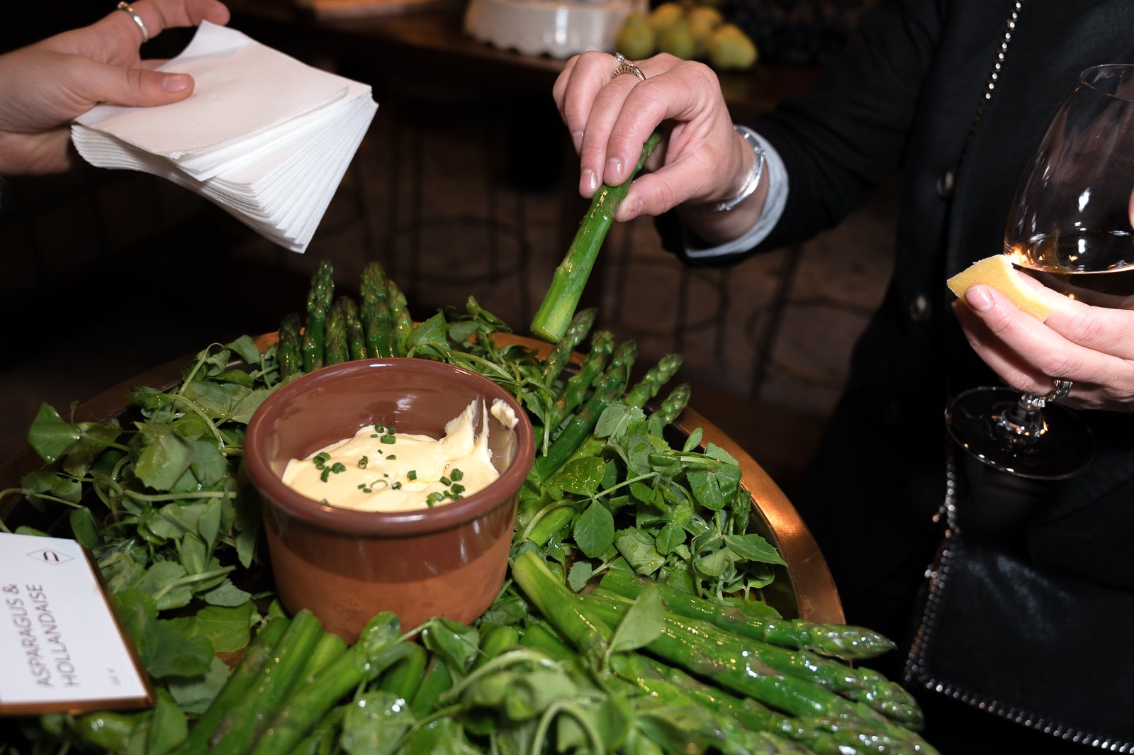 People preparing to dip fresh green asparagus in a sauce or dip at a table filled with asparagus and greens.
