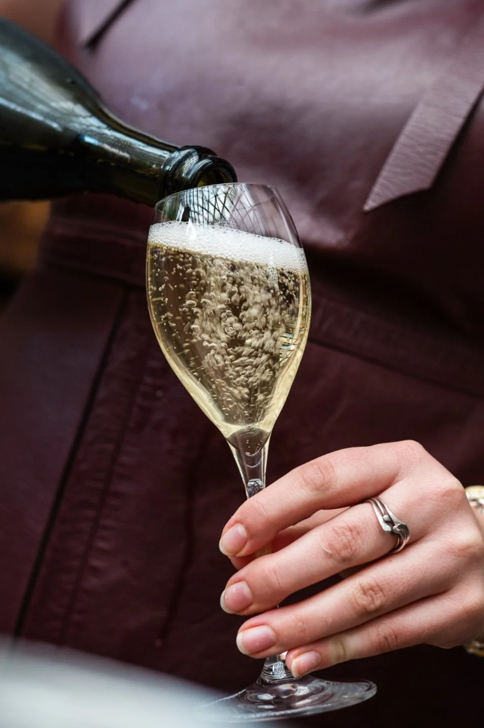A hand with a ring holding a champagne flute being filled with sparkling champagne.