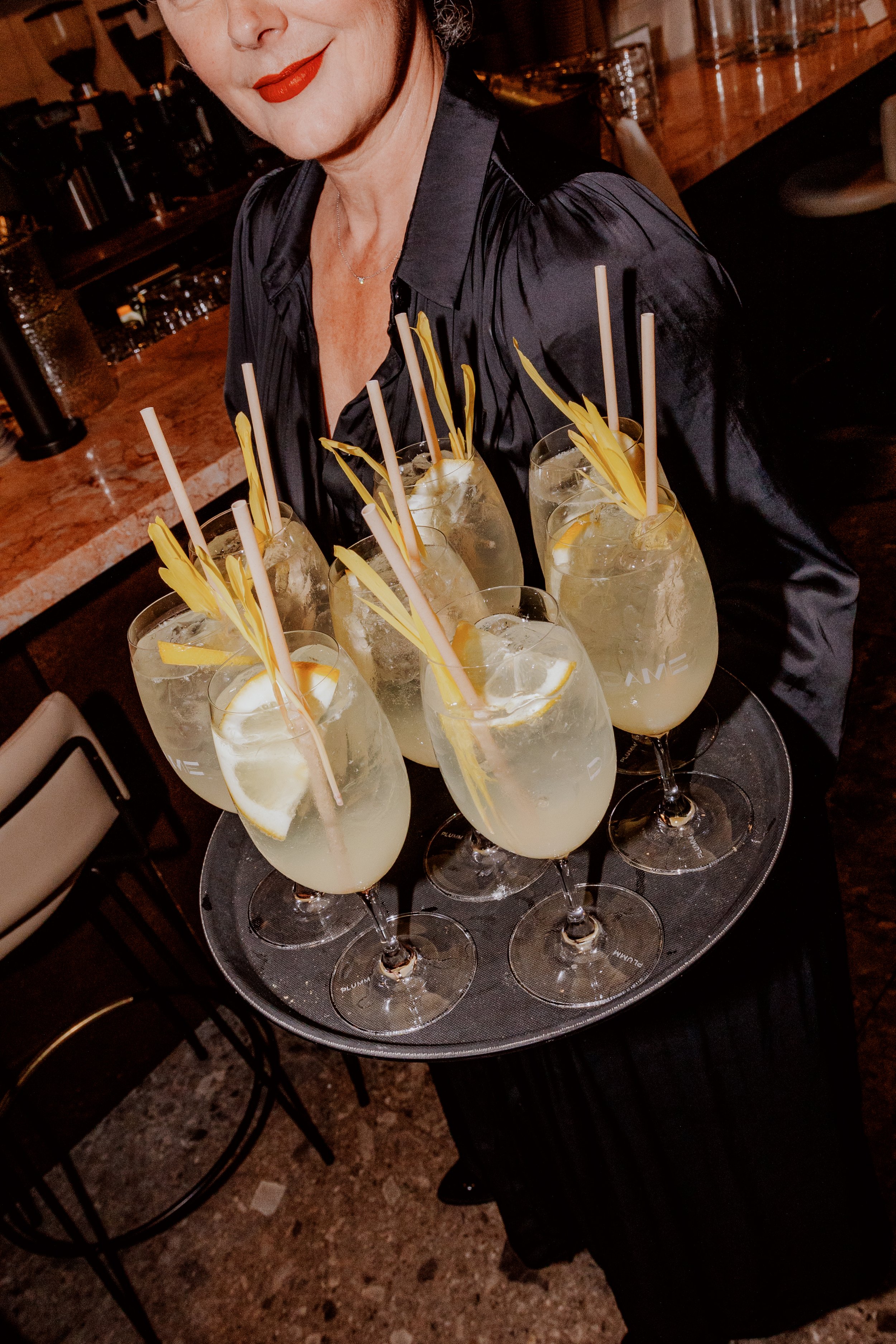 A woman with red lipstick and a black satin blouse holding a tray of large cocktails garnished with slices of lemon and decorated with yellow straw ornaments.