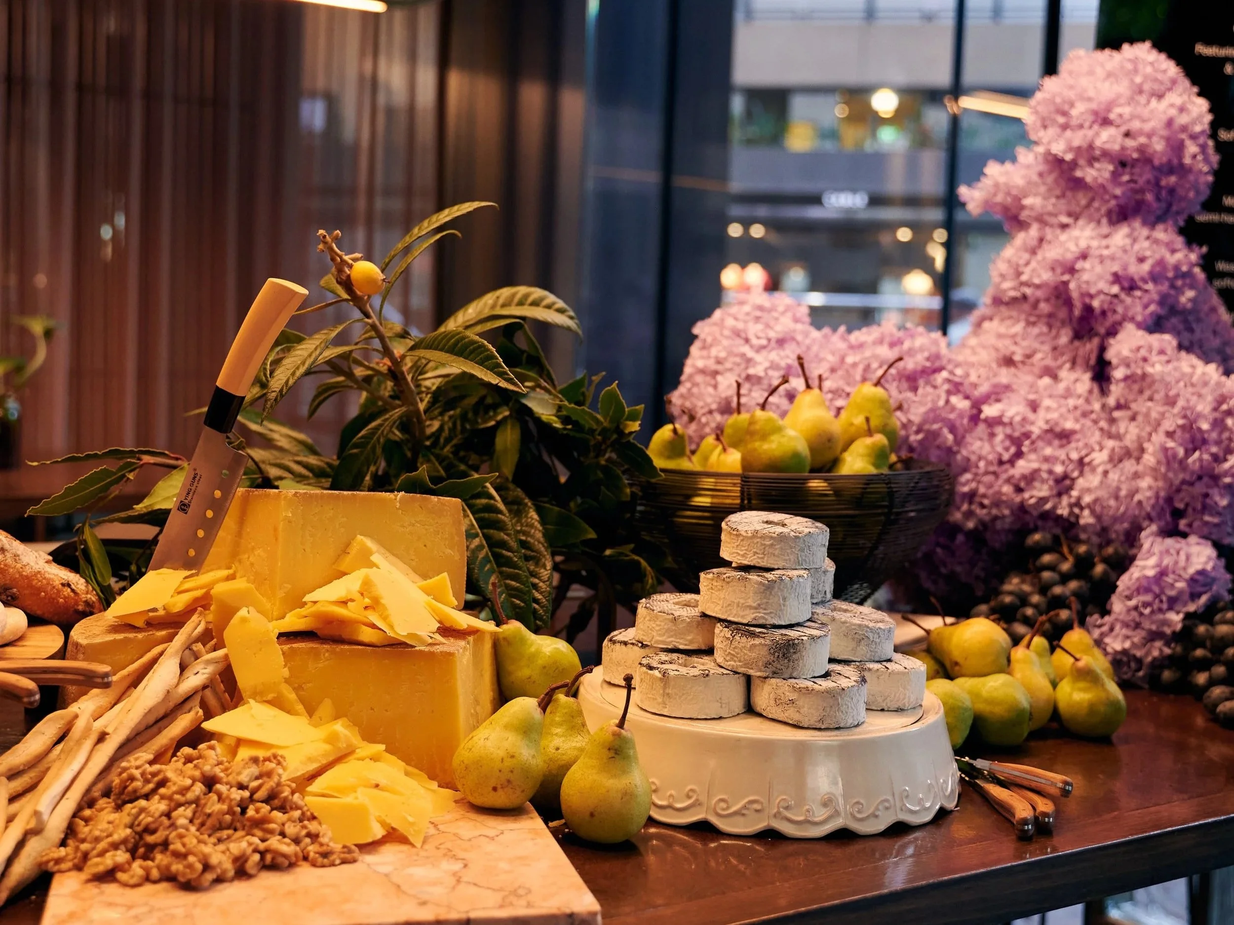 Display of cheese, pears, and other cheeses and snacks on a wooden table, with a background of flowers and pastries.