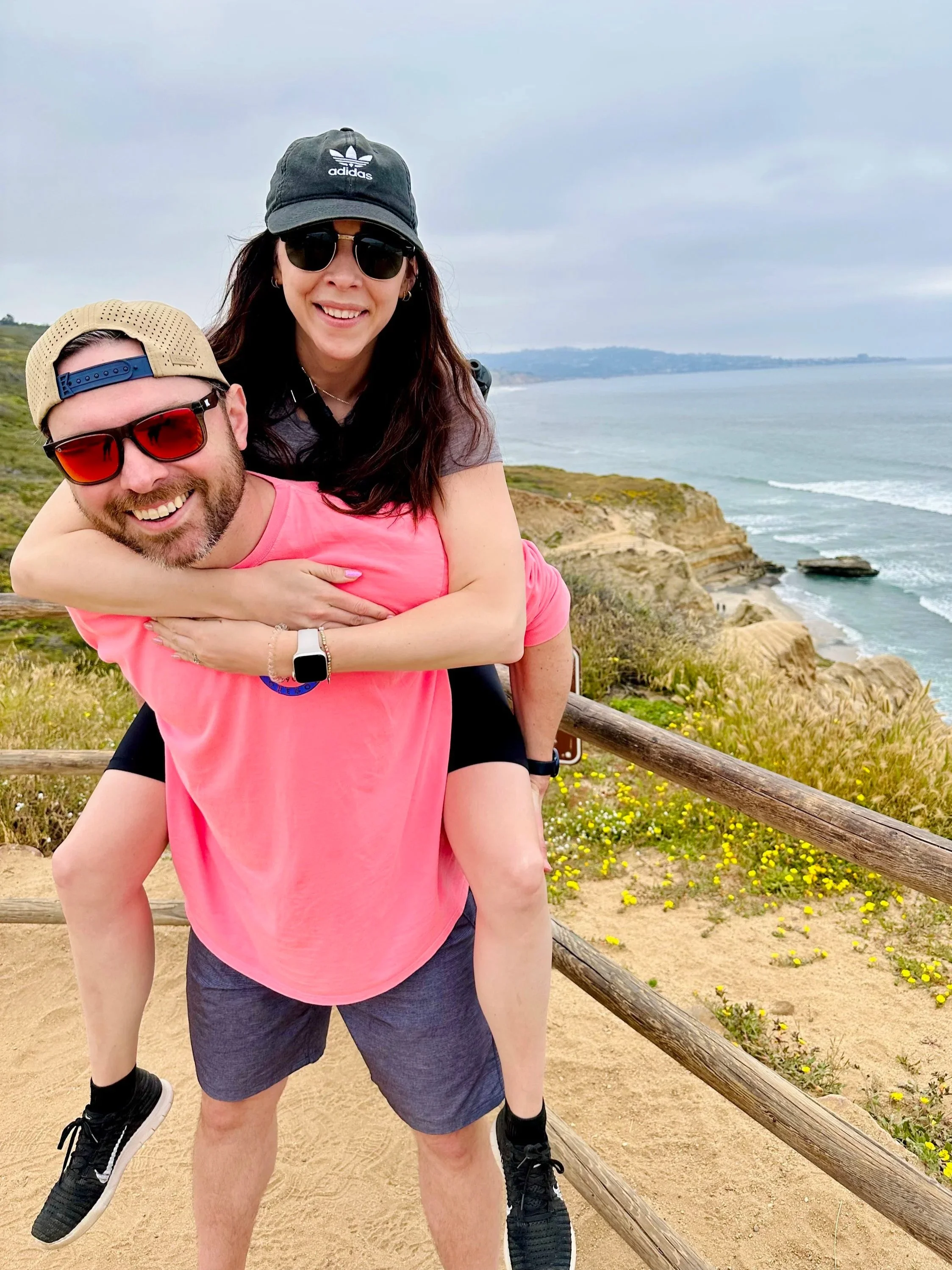 A man giving a piggyback ride to a woman on a coastline with cliffs, sandy beach, and ocean waves in the background.