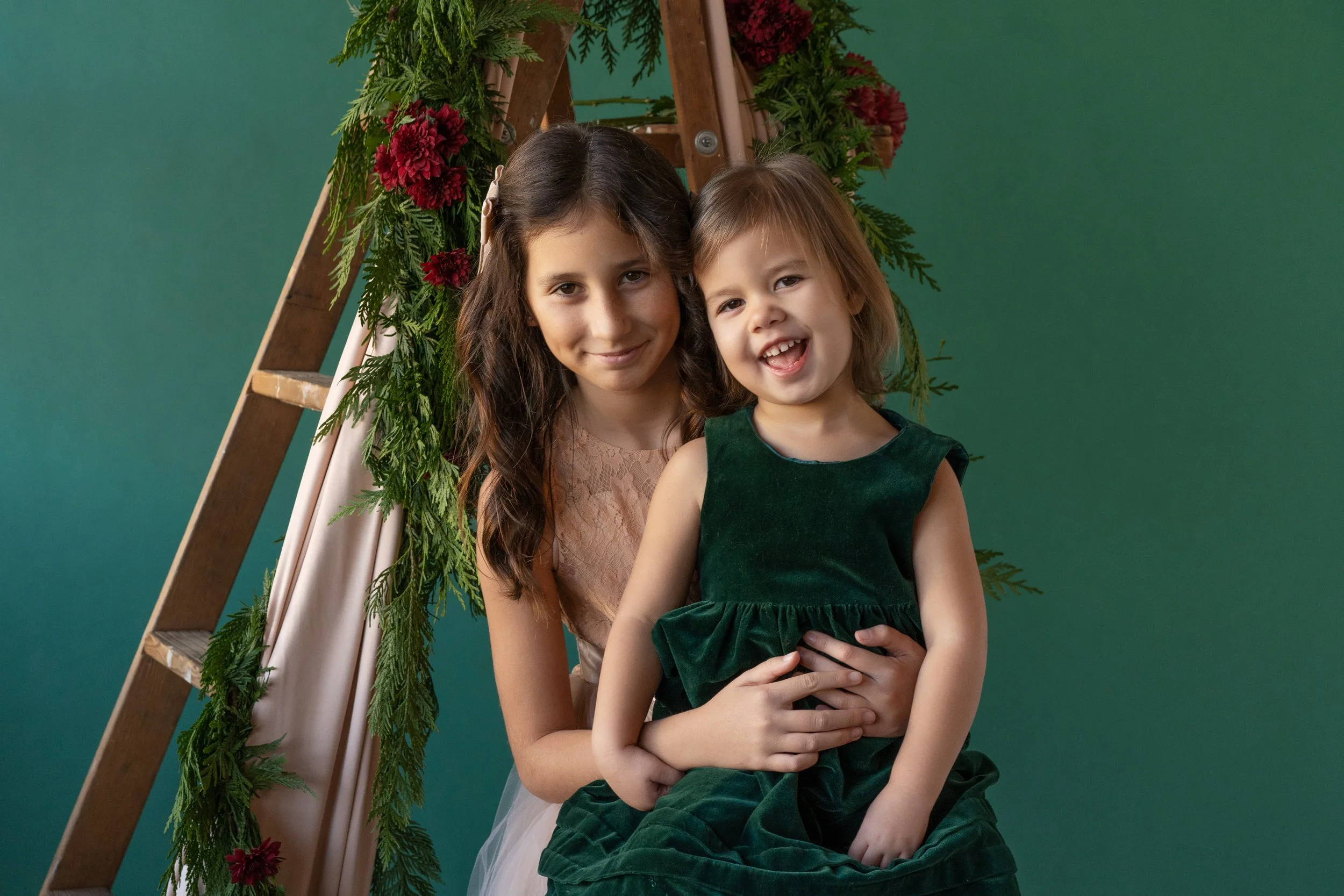 Two sisters dressed up on a green backdrop with garland and flowers behind them