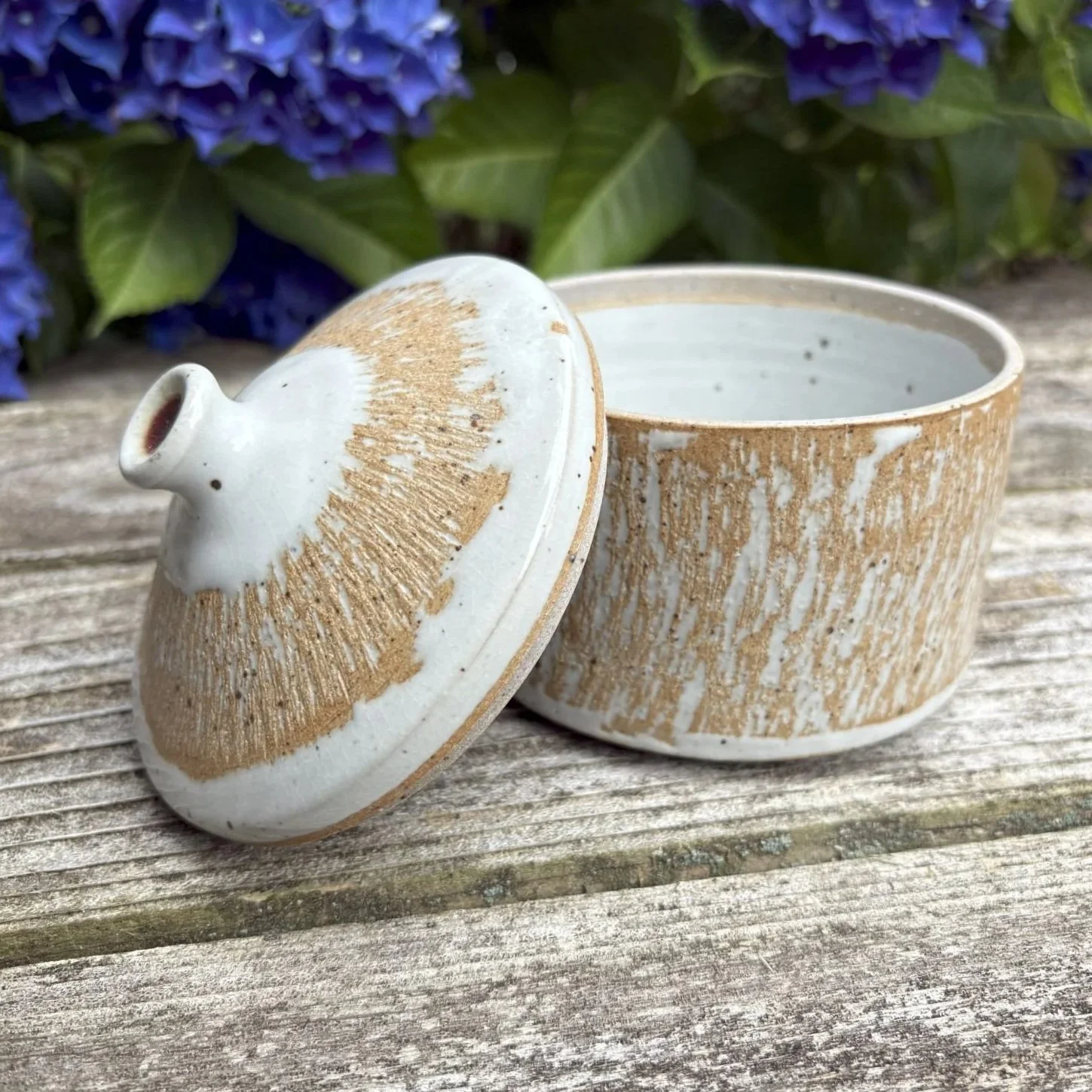 A ceramic container with chattered texture, glazed in white and featuring bare clay, with the lid slightly tilted to the side.