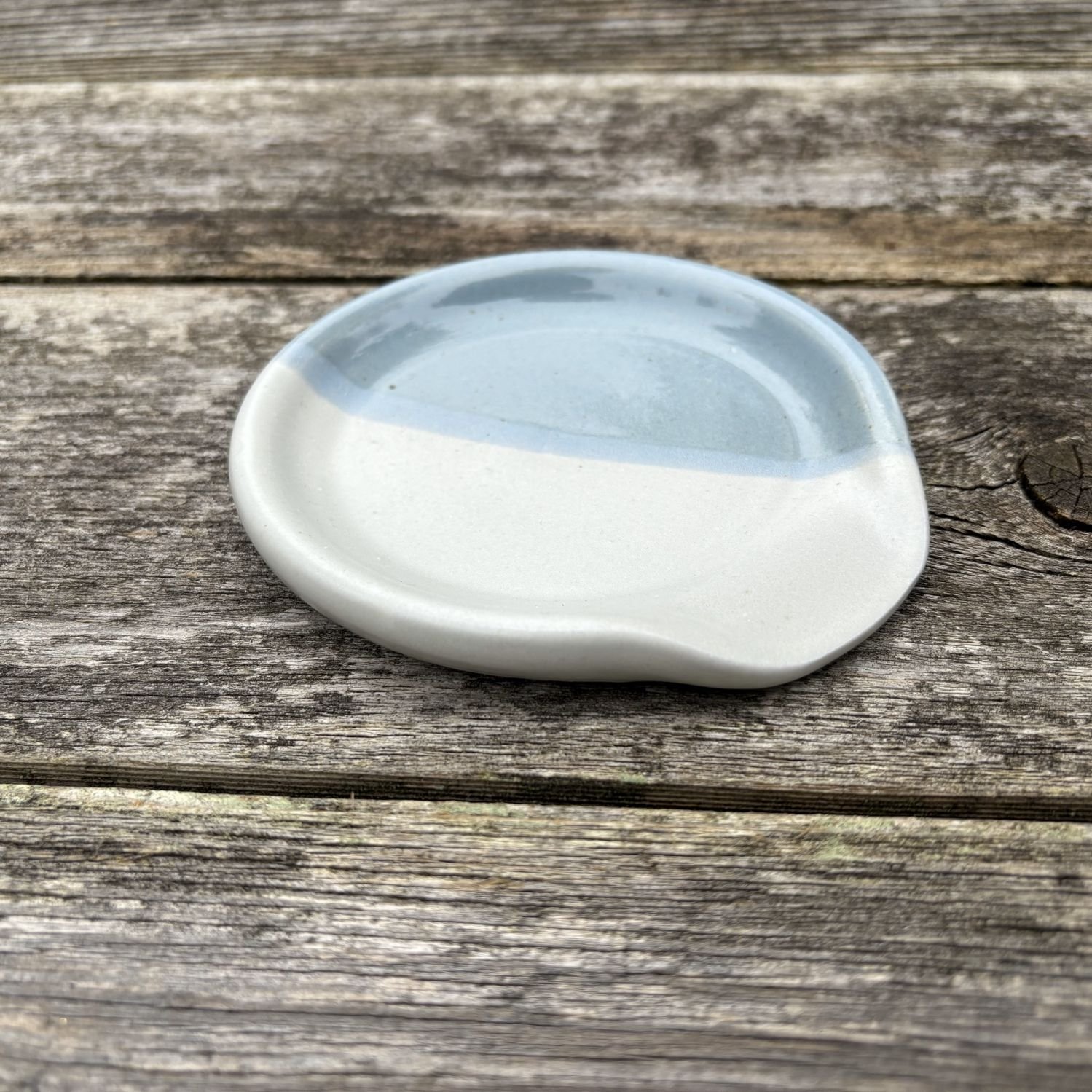 Side view of white and cornflower blue spoon rest placed on a rugged rock, highlighting the contrast between the dish and the aged fence..
