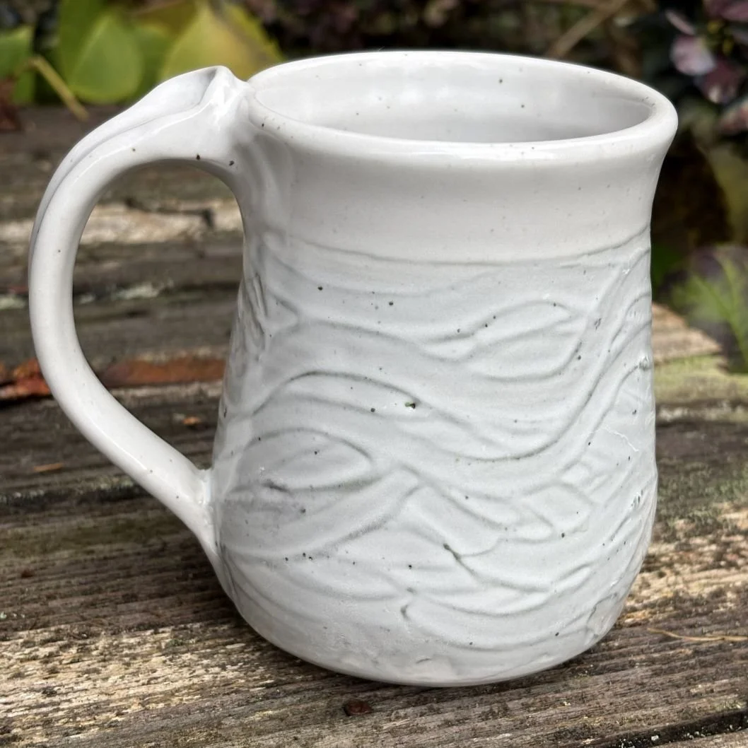 A white ceramic mug with a textured, wavy surface, sitting on a weathered wooden surface outdoors.