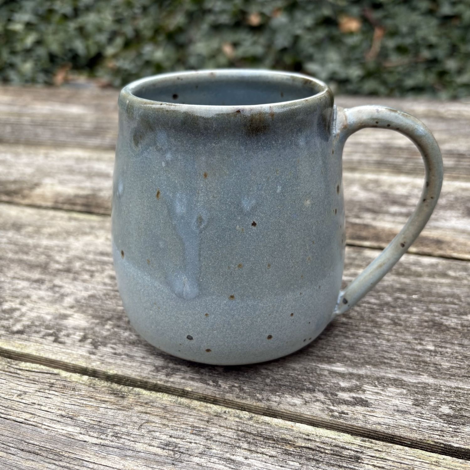 On rustic fence panel with greenery behind, mug with dark blue on the top half and light blue on the bottom half sits, with handle centered..