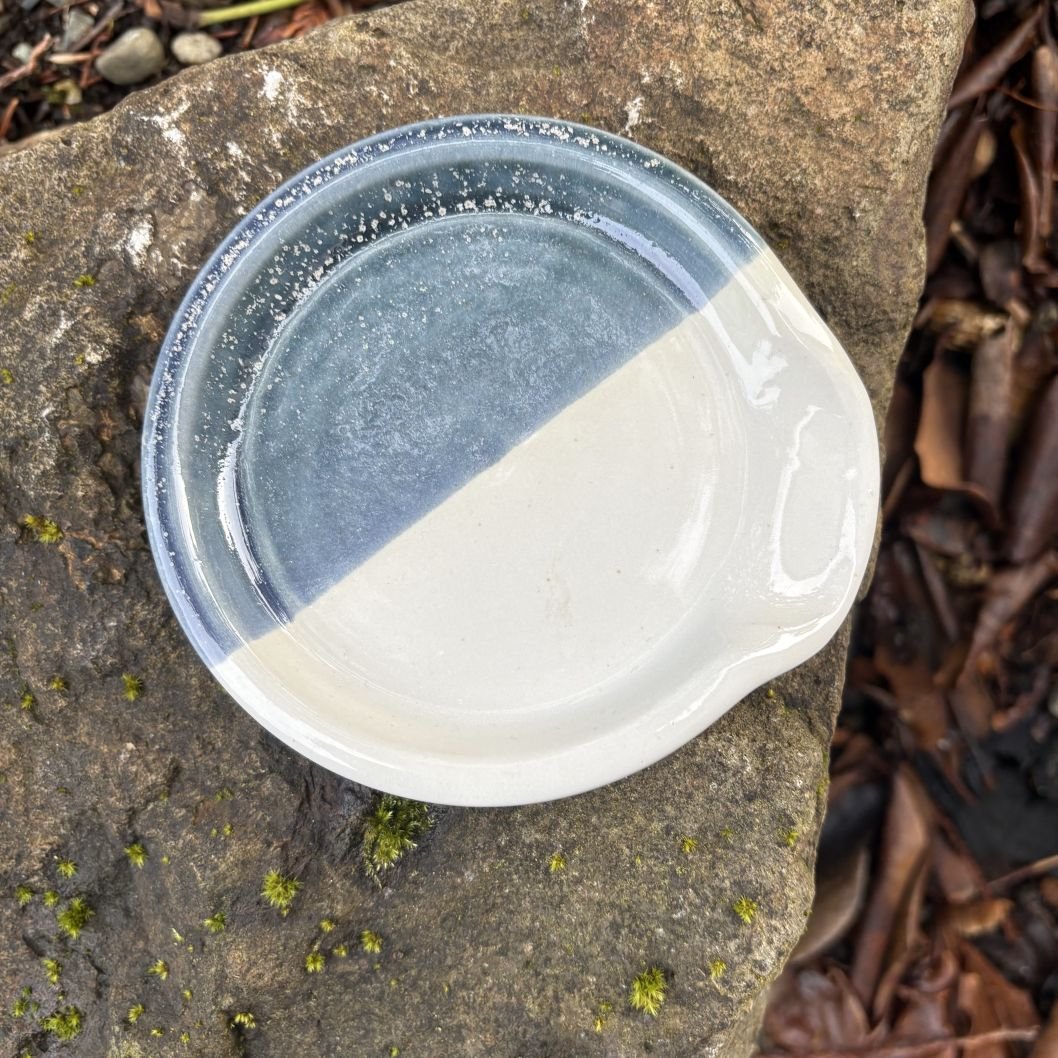 Overhead view of spoon rest glazed in shiny white and gray blue with white speckles placed on a rugged rock, highlighting the contrast between the dish and the natural stone.