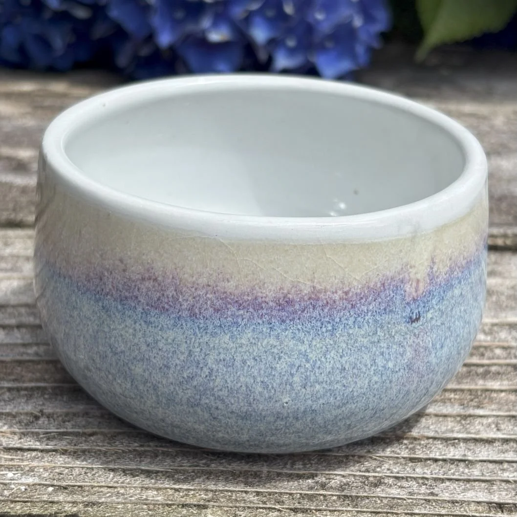 Ceramic bowl with a multicolored glaze, mainly shades of blue and purple, sitting on a weathered wooden surface with purple flowers in the background.