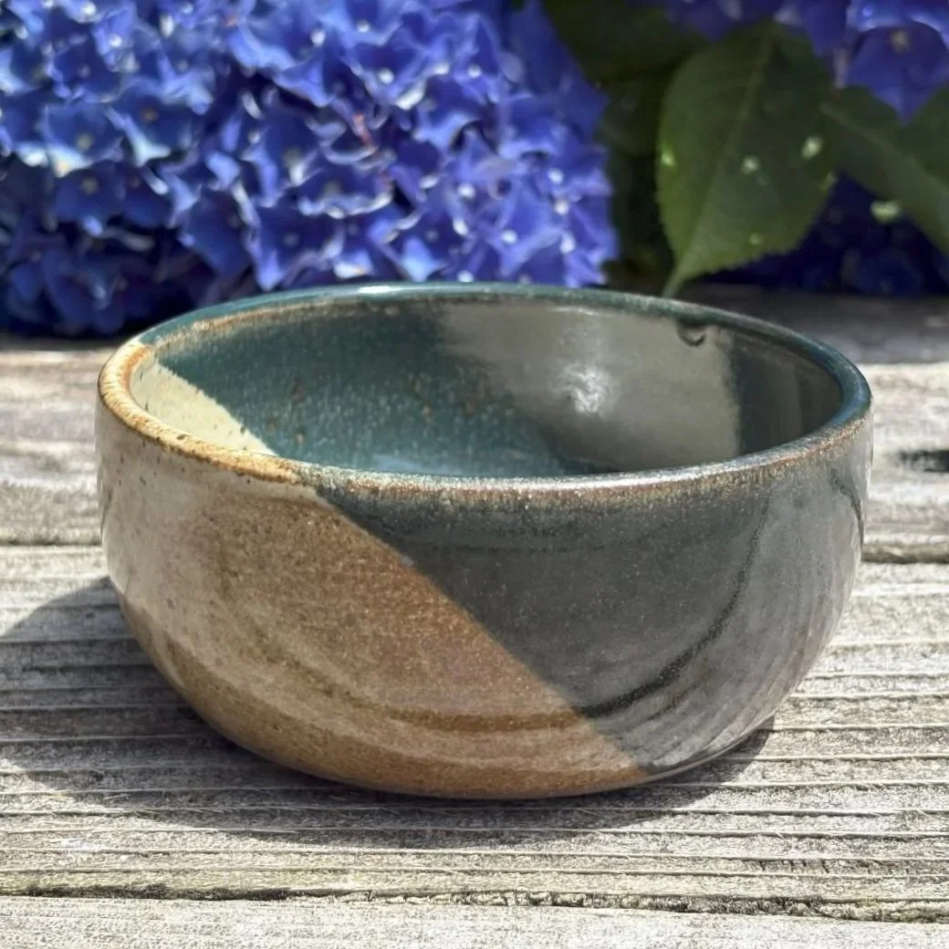 A small ceramic bowl with a multicolored glaze, sitting on a weathered wooden surface, with purple hydrangea flowers in the background.