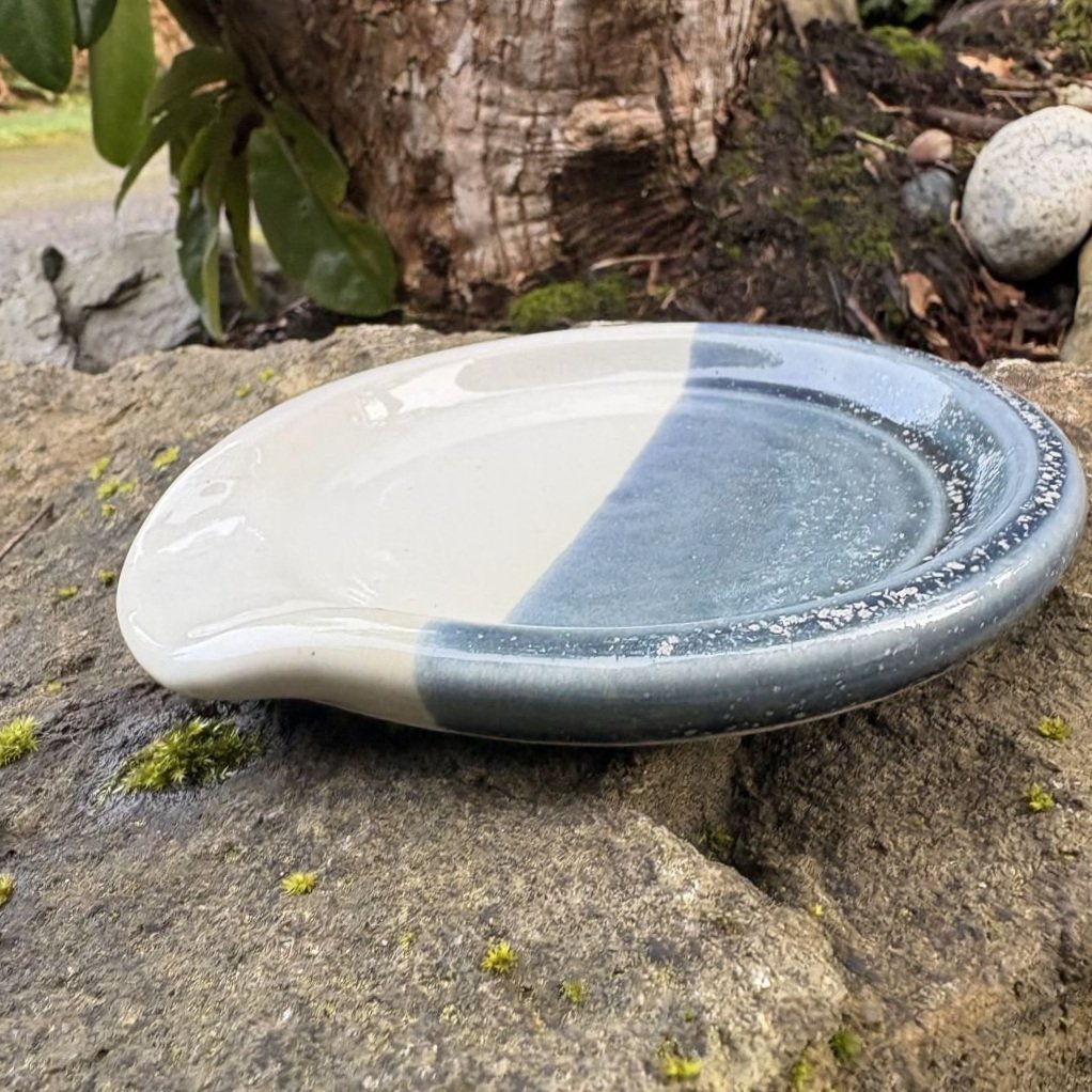 Side view of spoon rest glazed in shiny white and gray blue with white speckles placed on a rugged rock, highlighting the contrast between the dish and the natural stone.