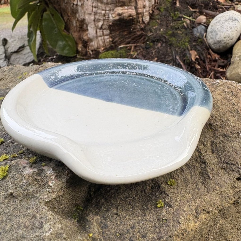 Side view of spoon rest glazed in shiny white and gray blue with white speckles placed on a rugged rock, highlighting the contrast between the dish and the natural stone.