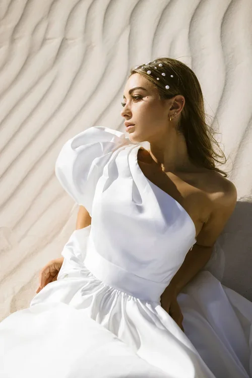 A woman in a white, off-the-shoulder Soleil dress with puffed sleeves sits on rippled sand, wearing a pearl-studded headband and gold hoop earrings, gazing thoughtfully into the distance.