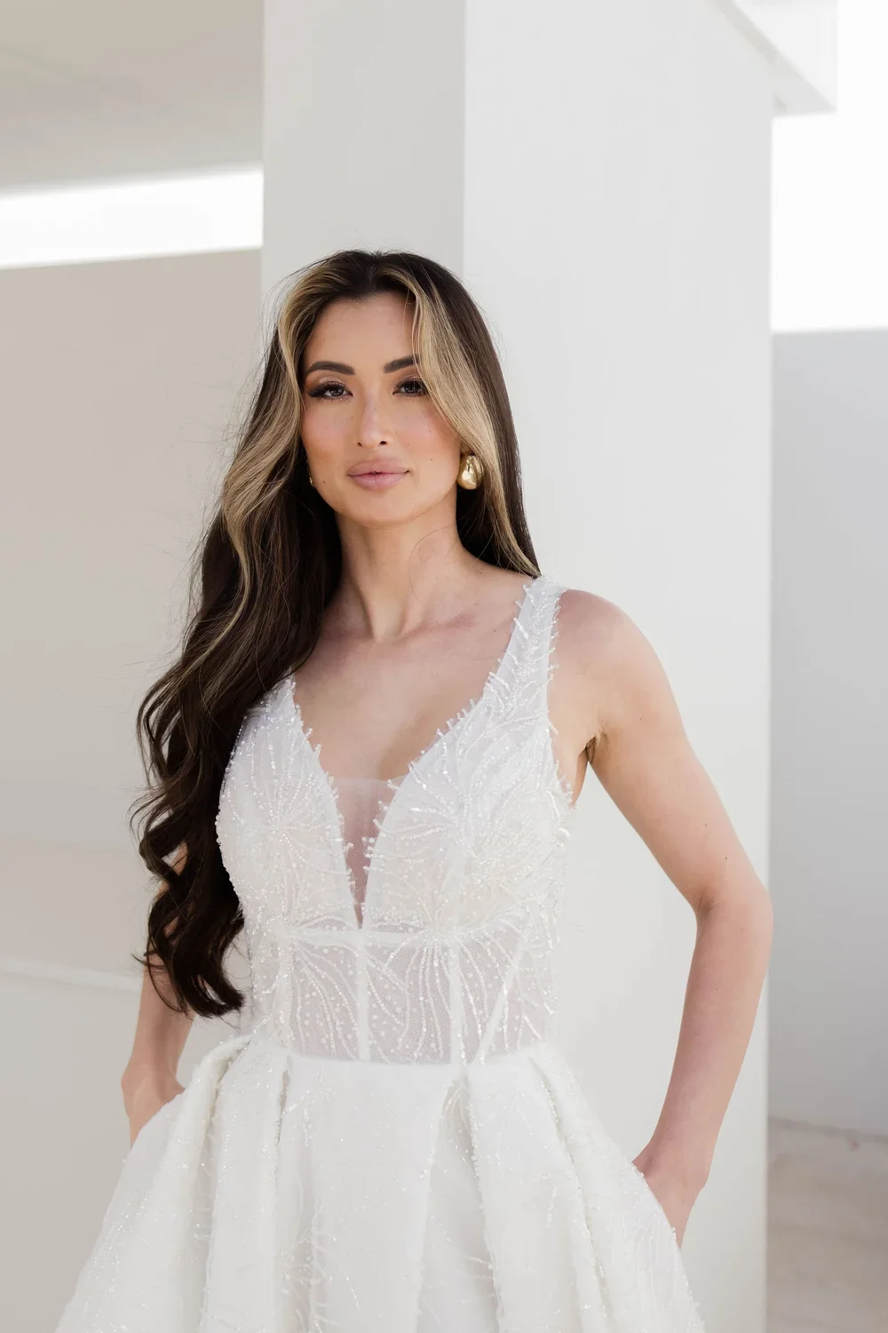 A woman with long, wavy hair and gold earrings wearing a ZC22824 sleeveless, white, beaded gown with a deep V neckline. She stands confidently in a bright, minimalist setting.