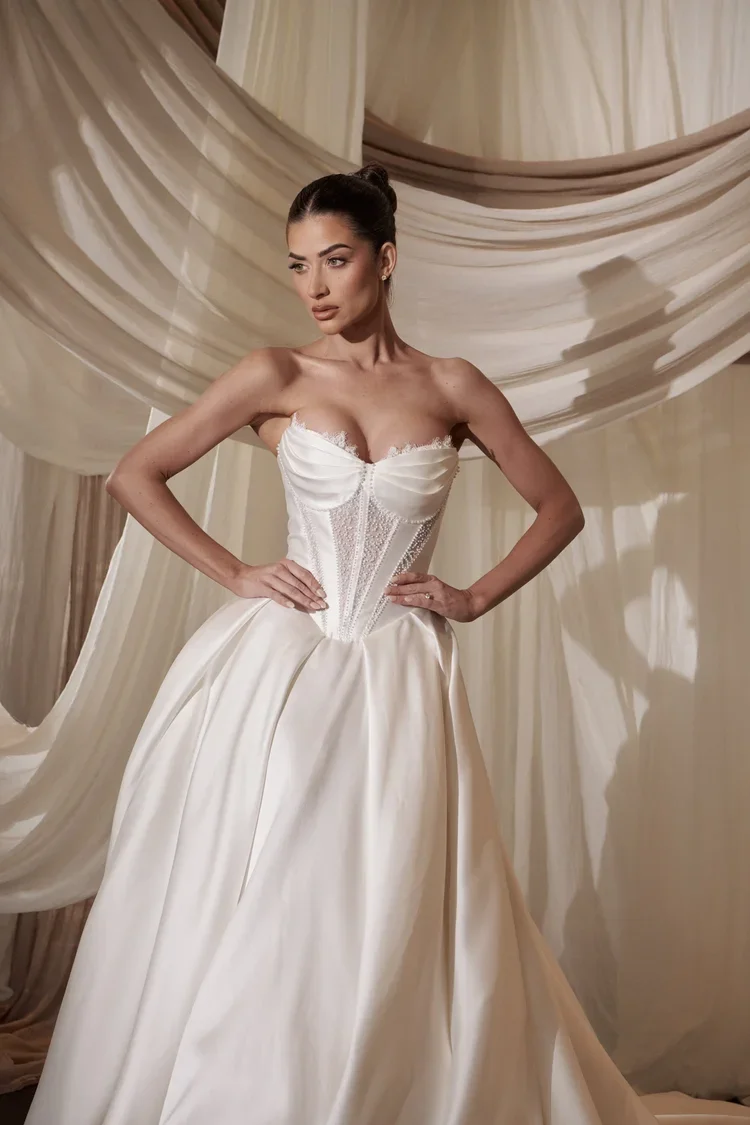 A woman stands with hands on hips, wearing a strapless white  Valencia wedding gown with a structured corset bodice and full skirt. Elegant draped fabric hangs in the background, creating a soft, romantic atmosphere.