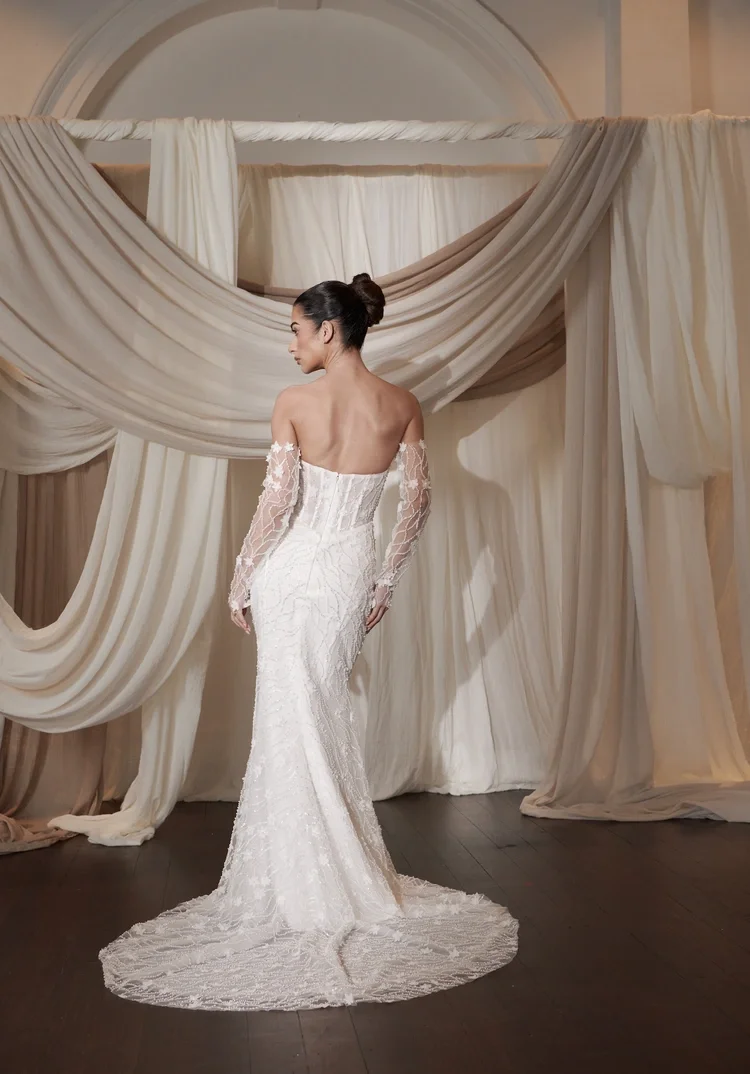A woman in an off-shoulder, long-sleeved, white lace wedding gown stands with her back to the camera in front of draped cream and beige fabric.