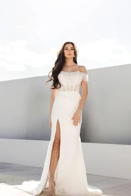 A woman in a white Serene off-the-shoulder wedding dress with a sheer lace bodice and high slit stands outdoors on a sunny day, with long wavy hair and neutral heels.