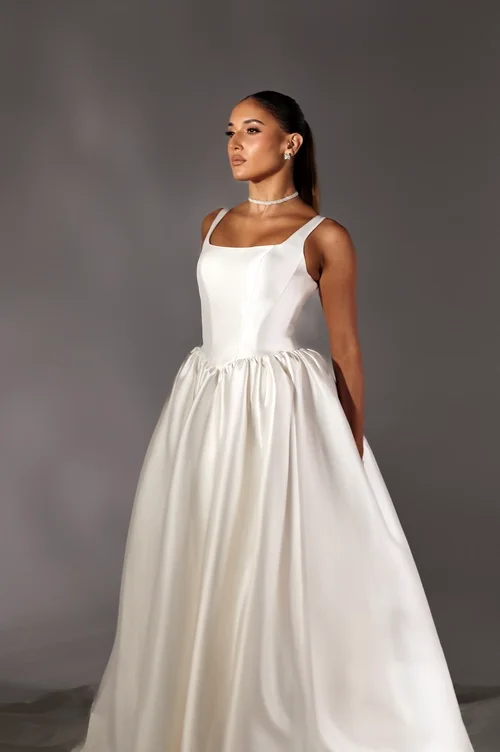 A woman wearing a sleeveless, white Milana ball gown with a fitted bodice and a flared skirt stands against a plain gray background. She has her hair pulled back and is accessorized with a pearl necklace and earrings.