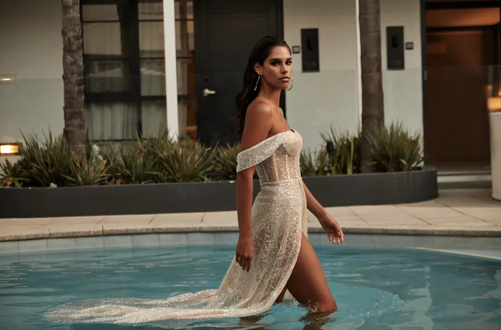 A woman in a sparkling, off-shoulder, sheer gown with a thigh-high slit walks through a shallow pool, with modern buildings and greenery in the background.