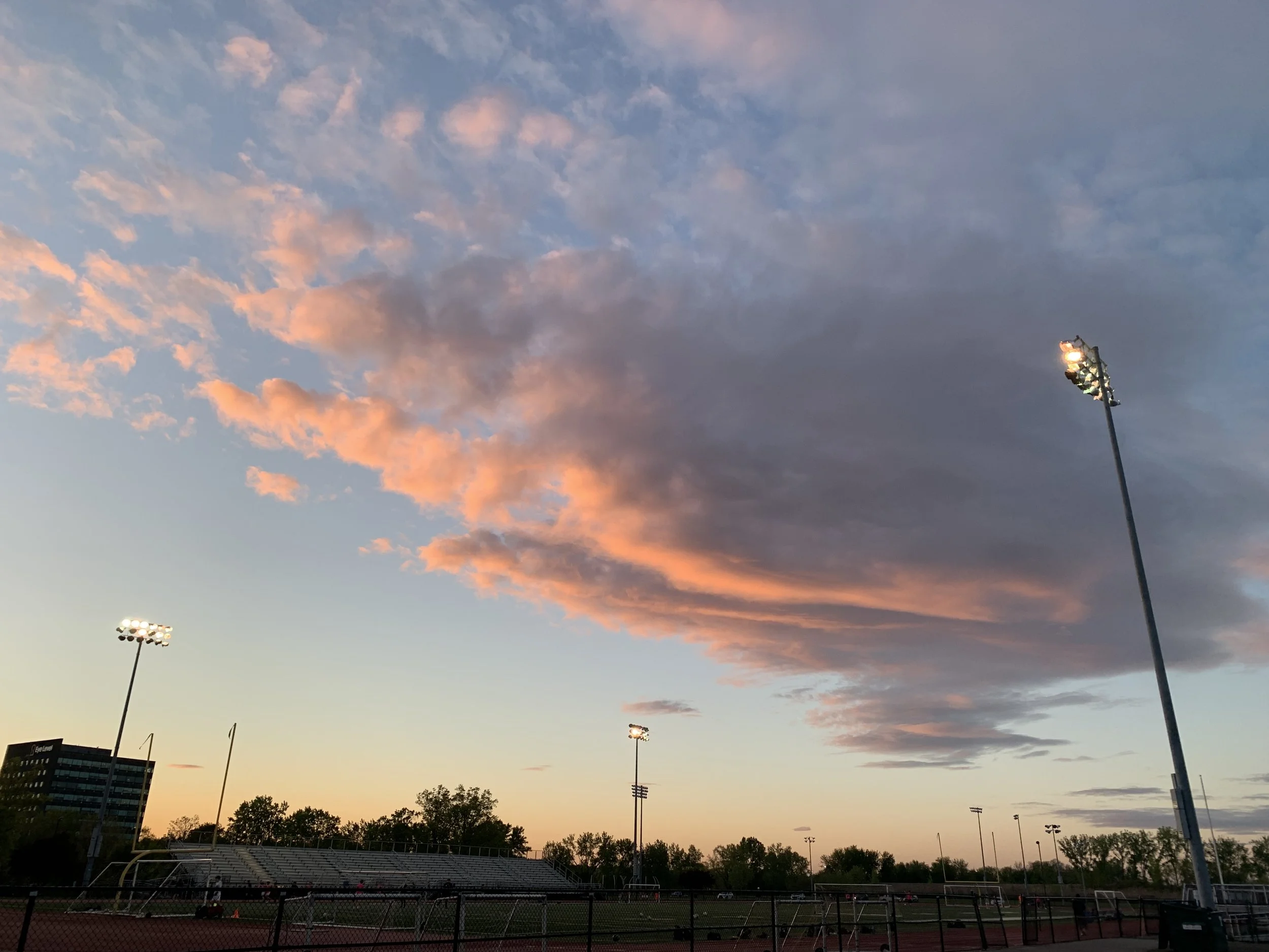 View of a sports field during sunset with pink and purple clouds in the sky, illuminated floodlights, and empty bleachers.