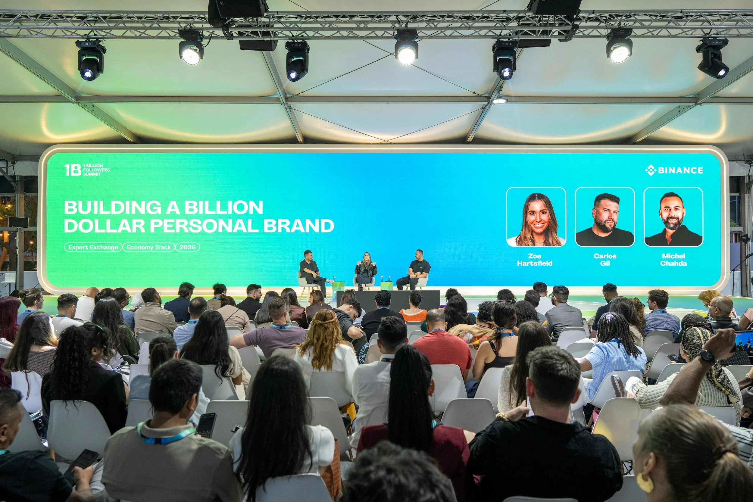 A conference event with a large digital screen displaying text about building a billion dollar personal brand, with three speakers seated on stage and a diverse audience listening.