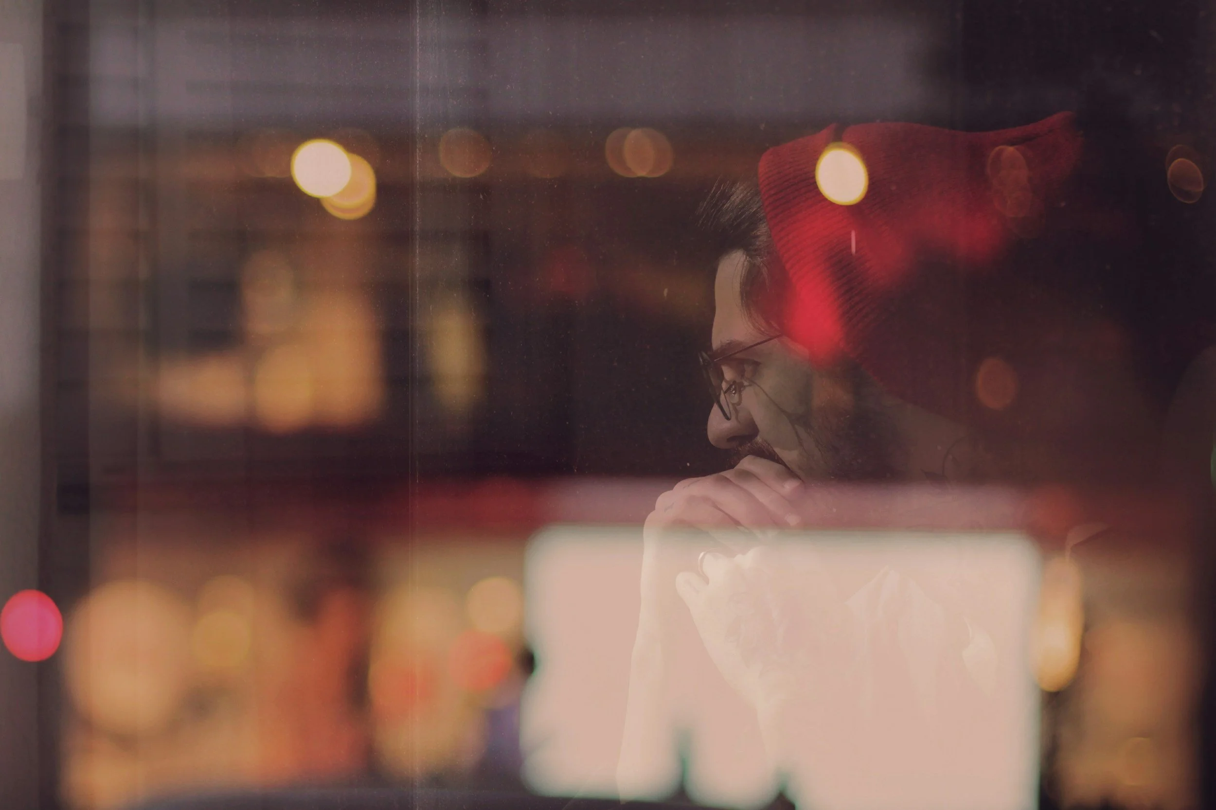 A woman with curly hair and glasses wearing a red beanie, resting her chin on her hand, seen through a window with blurred city lights in the background.