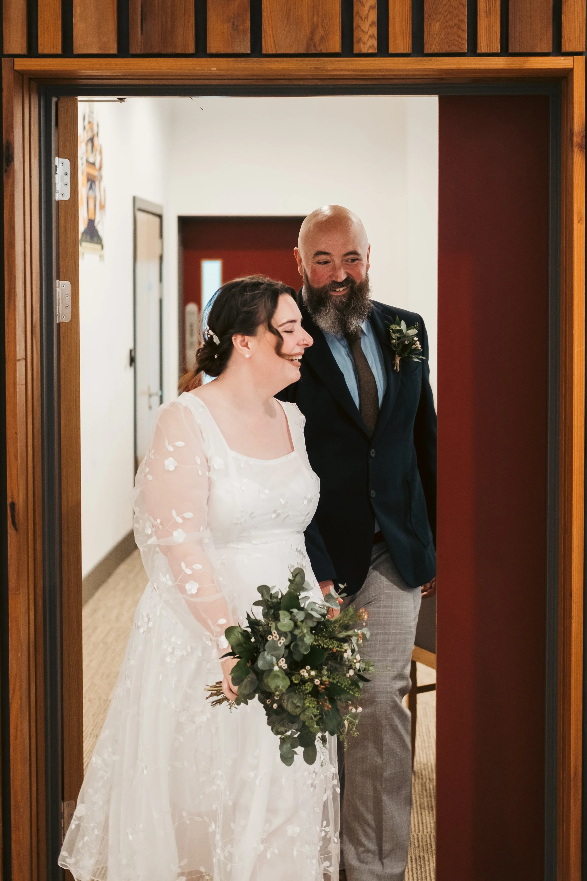 A bride and groom smiling and walking through a doorway indoors. The bride is holding a bouquet of greenery and flowers, wearing a white dress with floral embroidery. The groom is dressed in a dark blazer, light-colored pants, and a shirt with a tie,