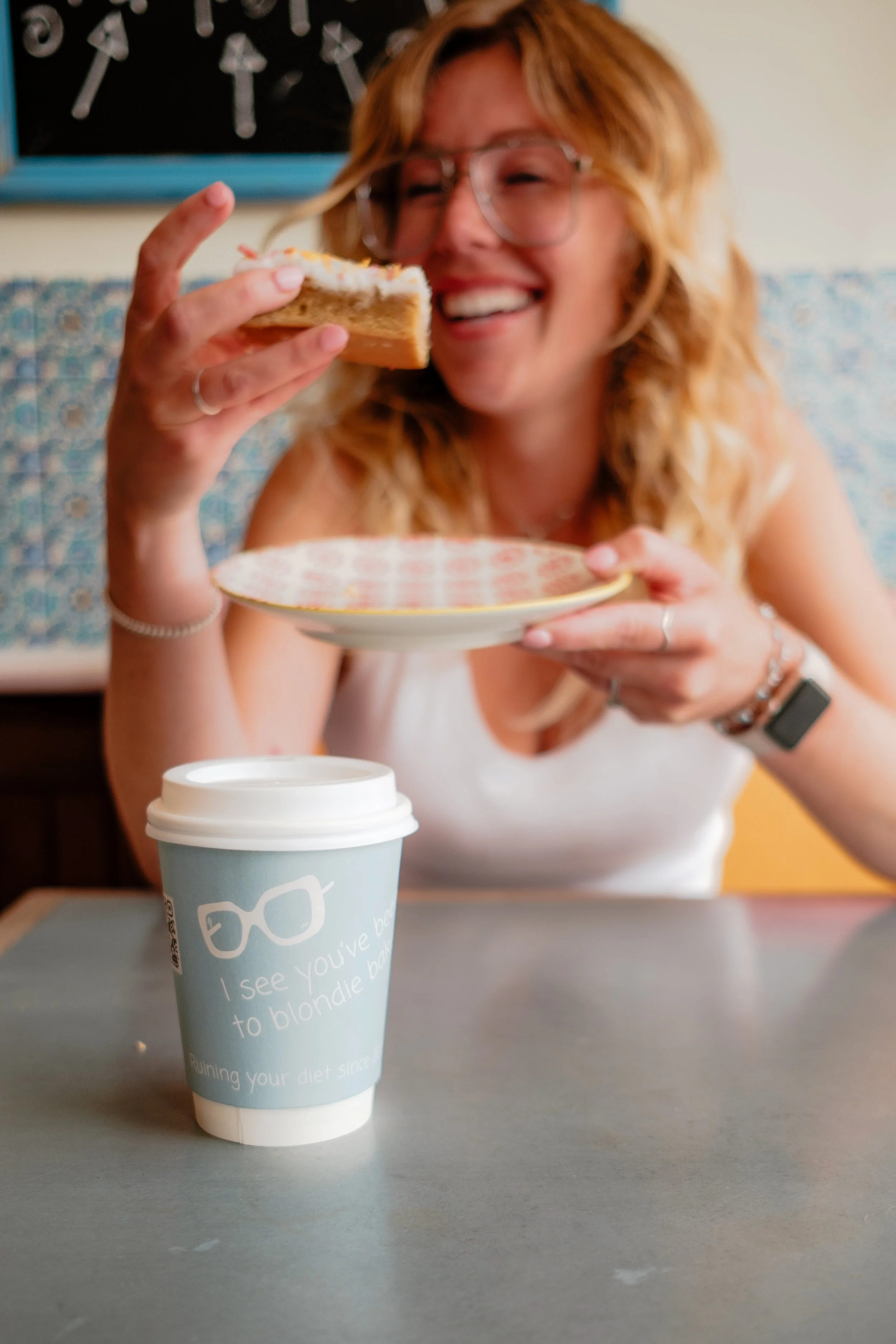 A woman with curly blonde hair, wearing glasses, smiling, holding a slice of cake and a plate. On the table, there is a blue disposable coffee cup with a lid.