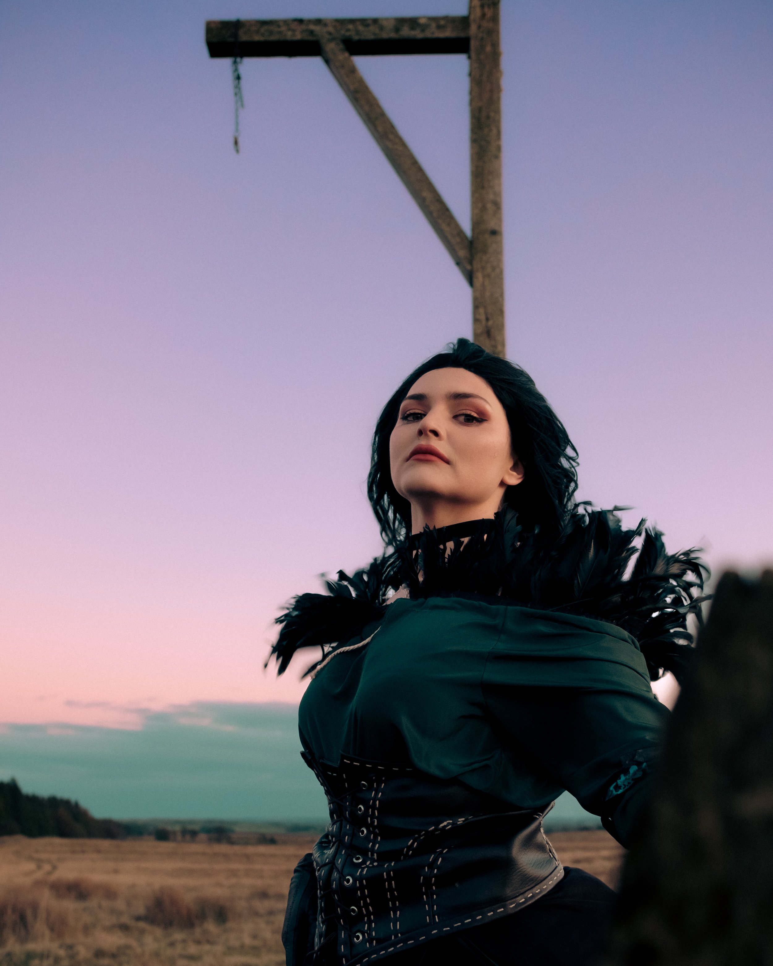 A woman with dark hair and makeup wearing a dark jacket and feathered accessory, standing outdoors during sunset with a sky in shades of pink and purple, a field, and hills in the background.