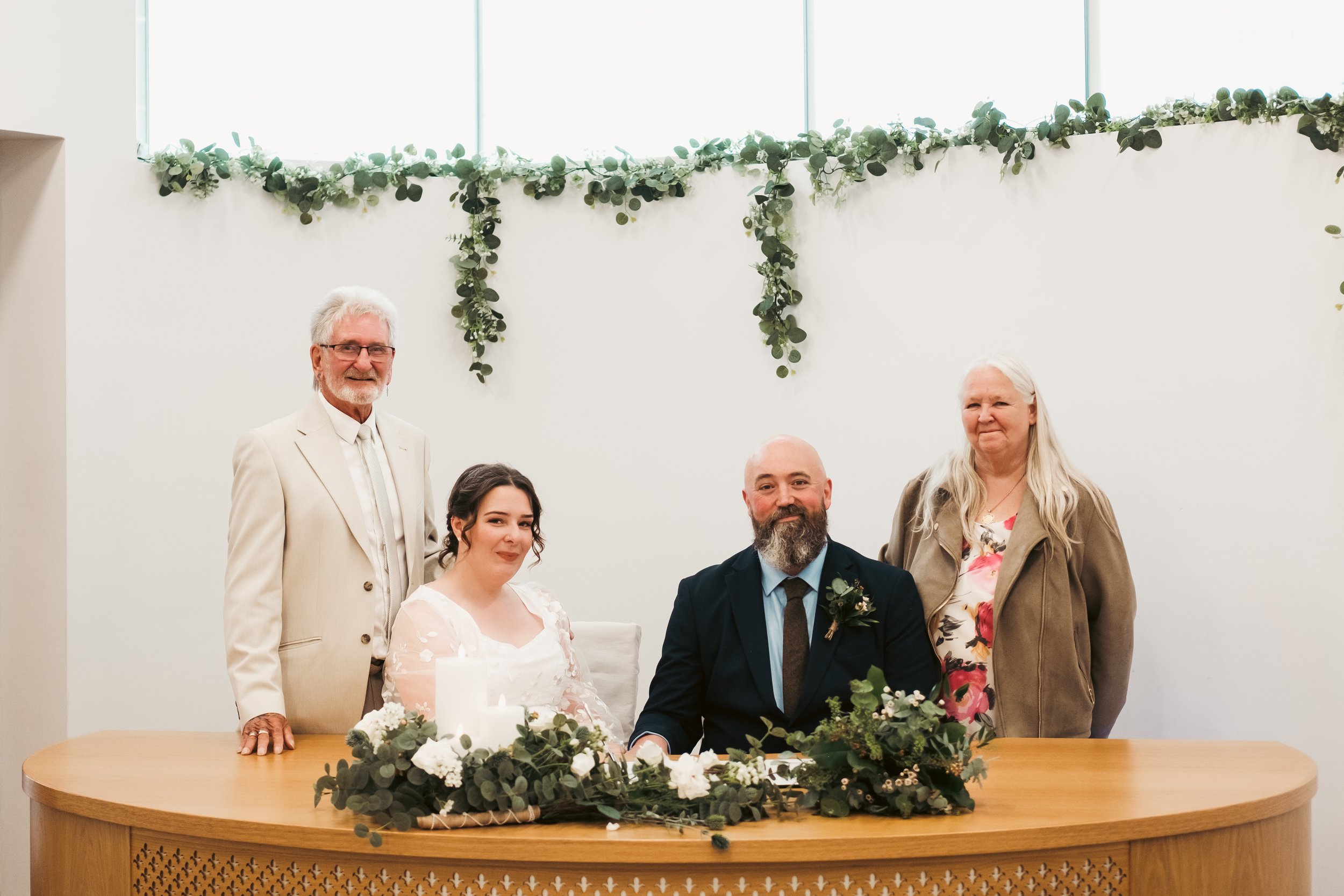 A wedding ceremony with five people, including a bride and groom seated at a table with floral decorations, and three standing individuals, inside a room with high windows and a green garland strip on the wall.