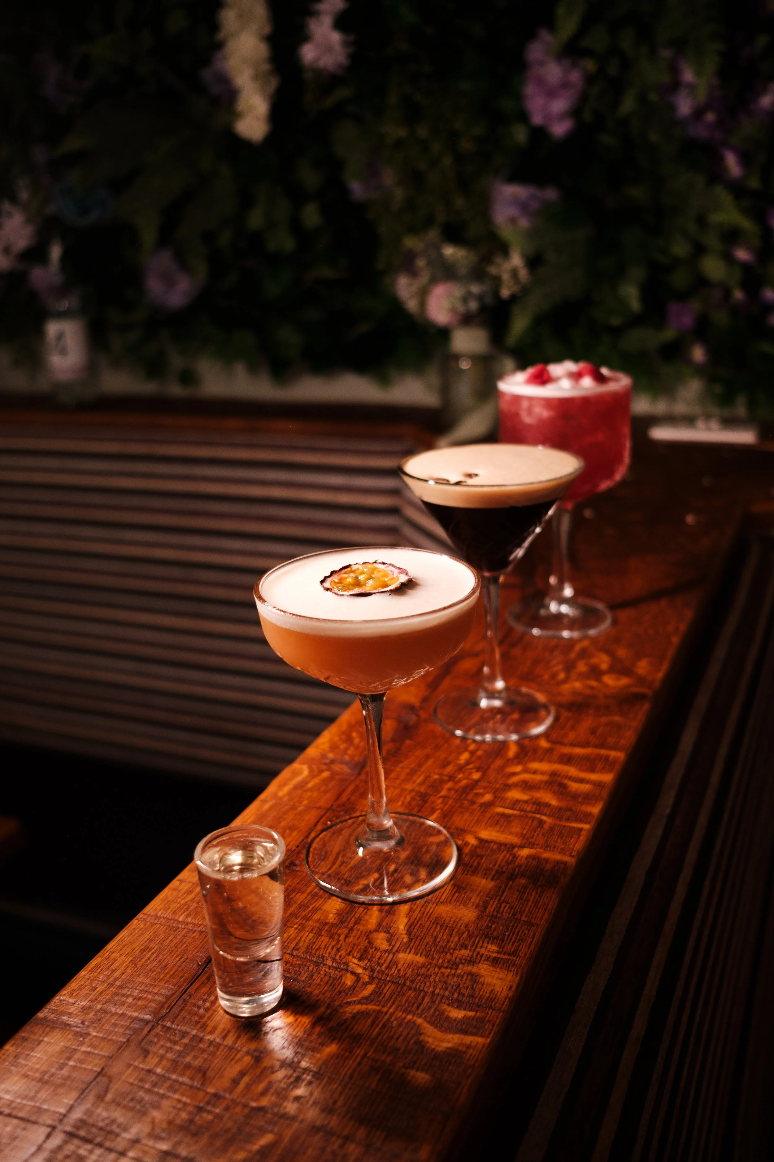 Three cocktails on a wooden bar counter with a floral background.