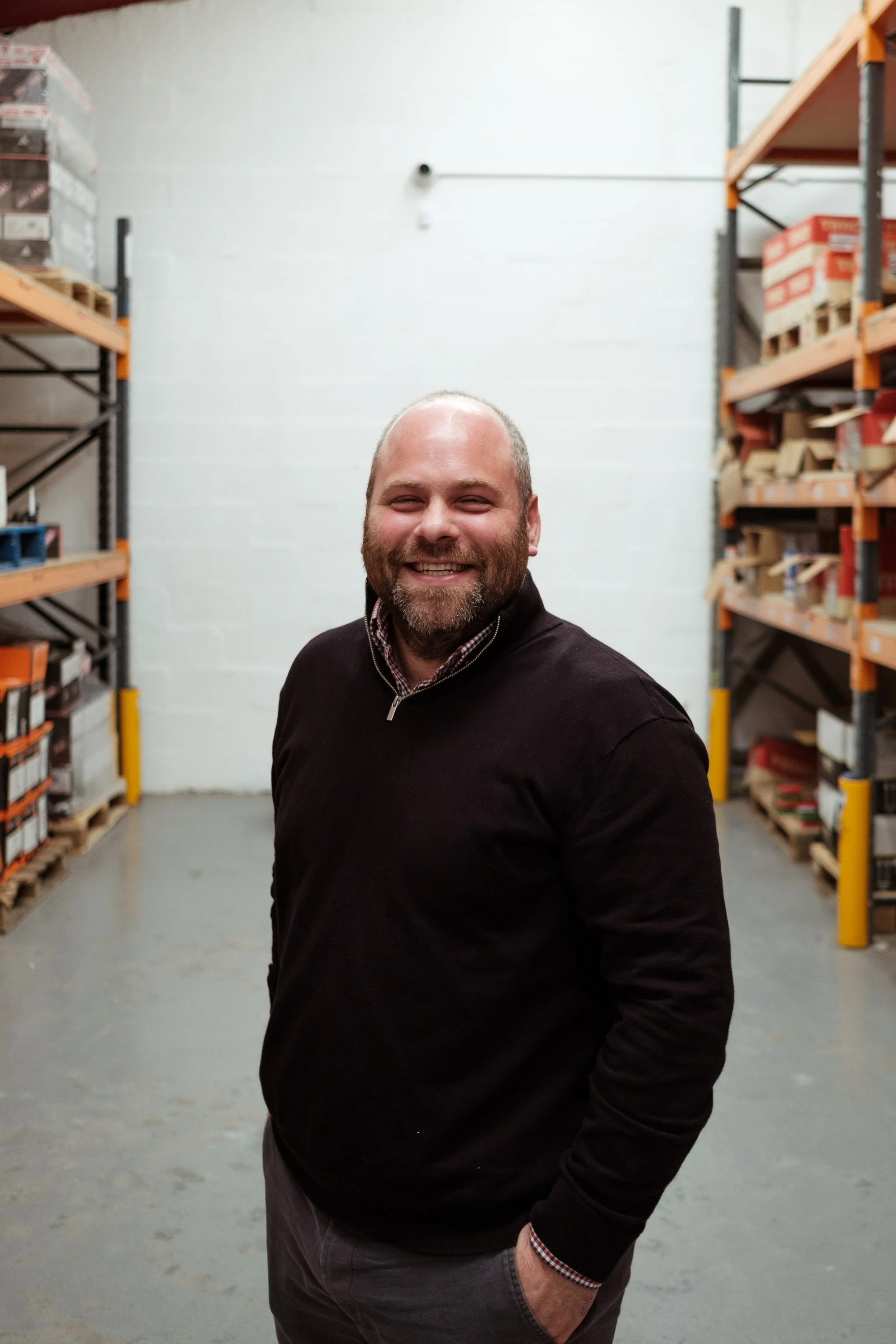 A smiling man with a beard standing in a warehouse aisle with shelves of boxes on either side.