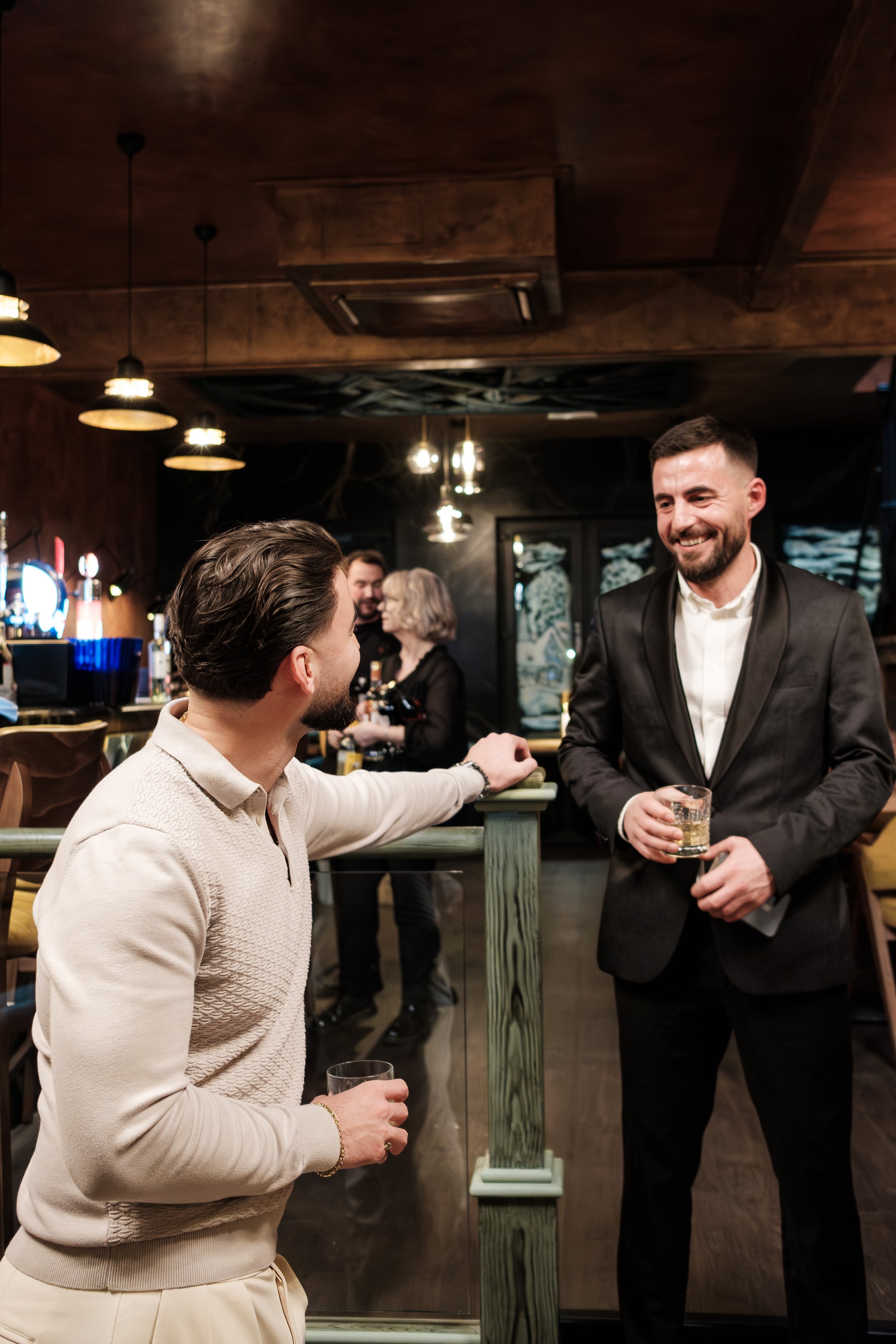 Two men at a bar talking and smiling, one holding a glass of whiskey, with other people in the background.