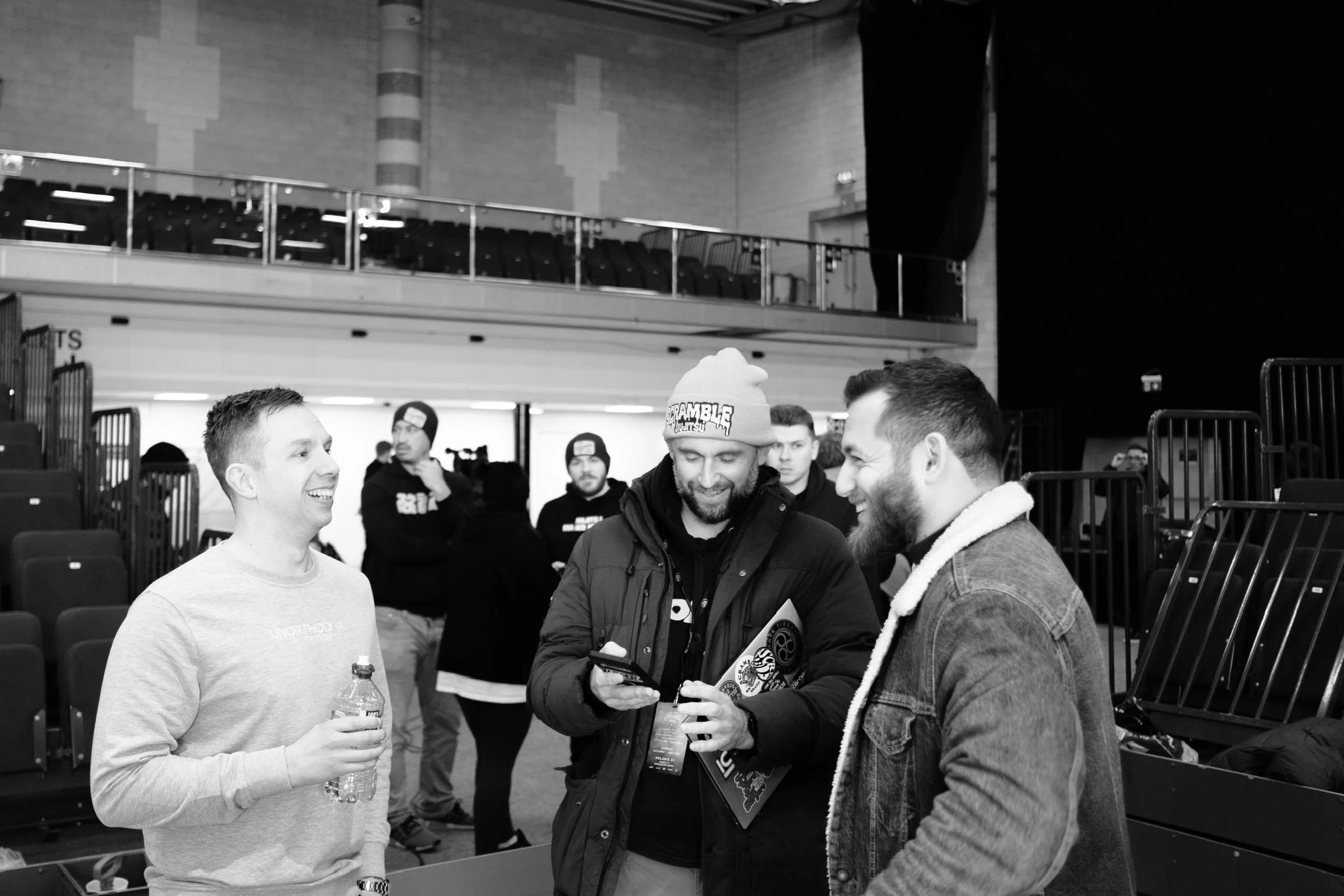 Three men are smiling and engaging in a conversation in an indoor arena or gymnasium setting. One man is holding a water bottle, another is looking at his phone, and the third is wearing a beanie. Several other people are in the background, some stan