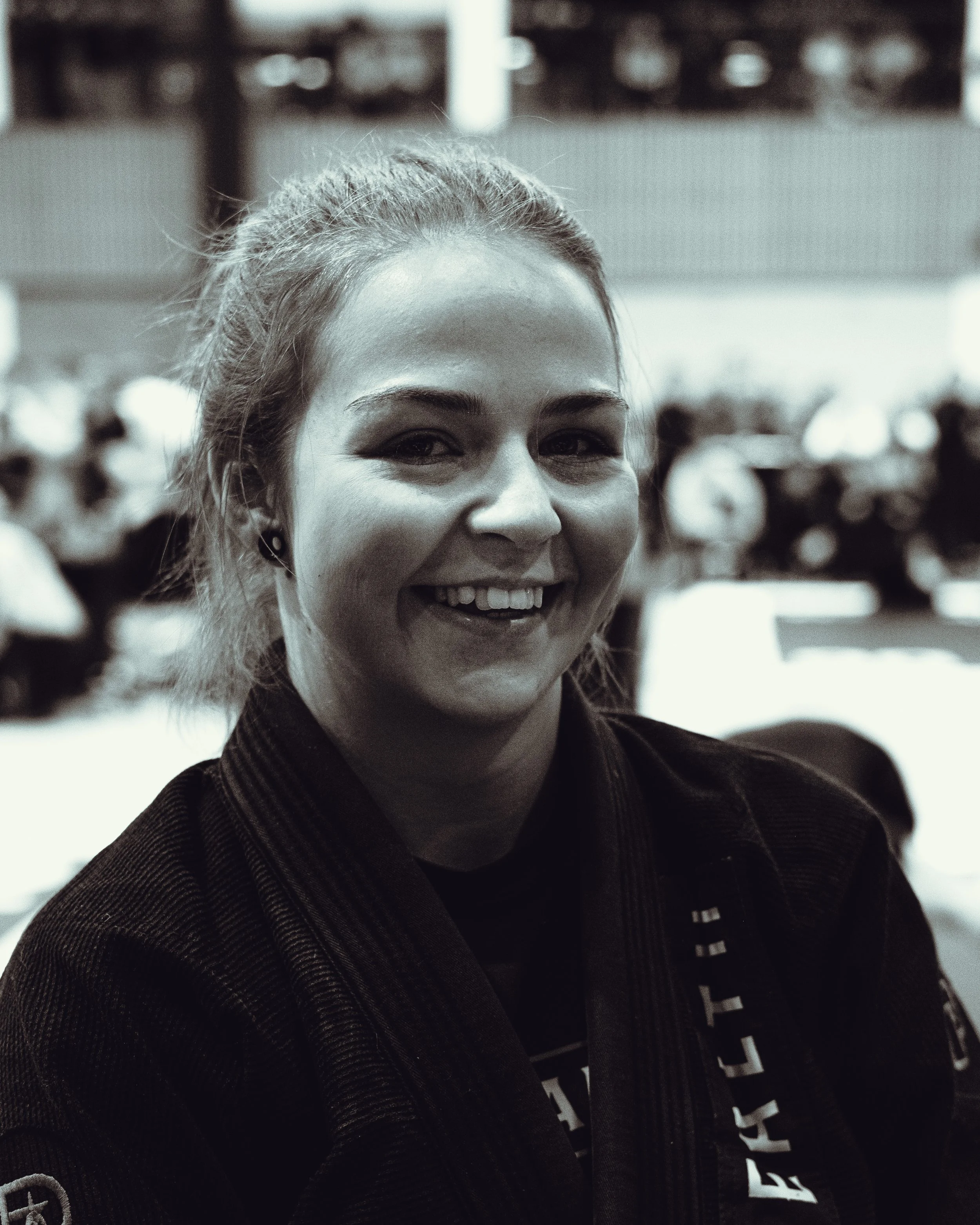 Close-up of a smiling woman with blonde hair tied back, wearing a black jacket, in a crowded indoor setting.