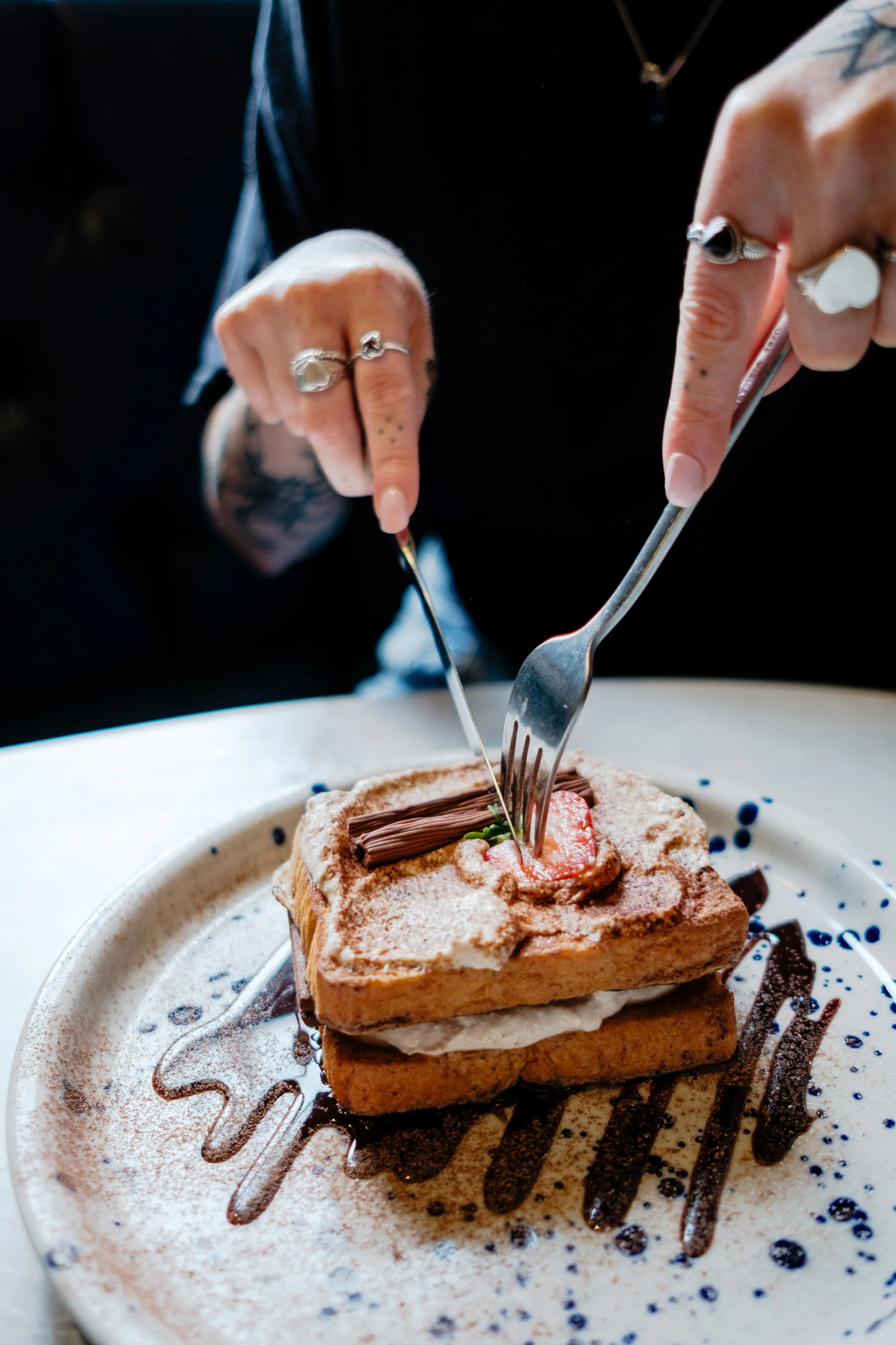 Person cutting into a layered dessert with a fork and knife, topped with chocolate and powdered sugar, on a plate decorated with chocolate drizzle.
