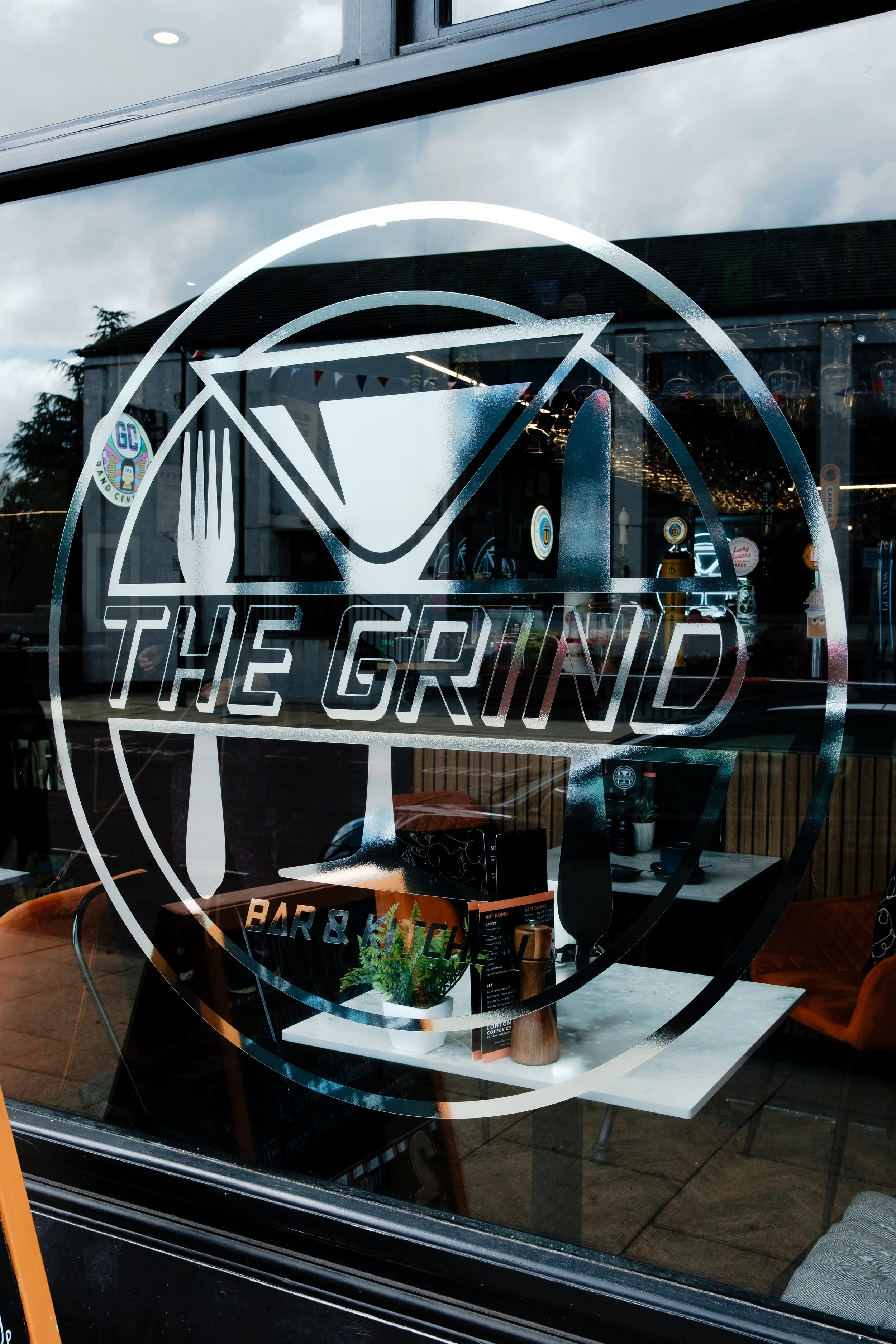 Glass window of a restaurant named 'The Grind' with a white logo depicting a martini glass, a fork, and a knife inside a circle.