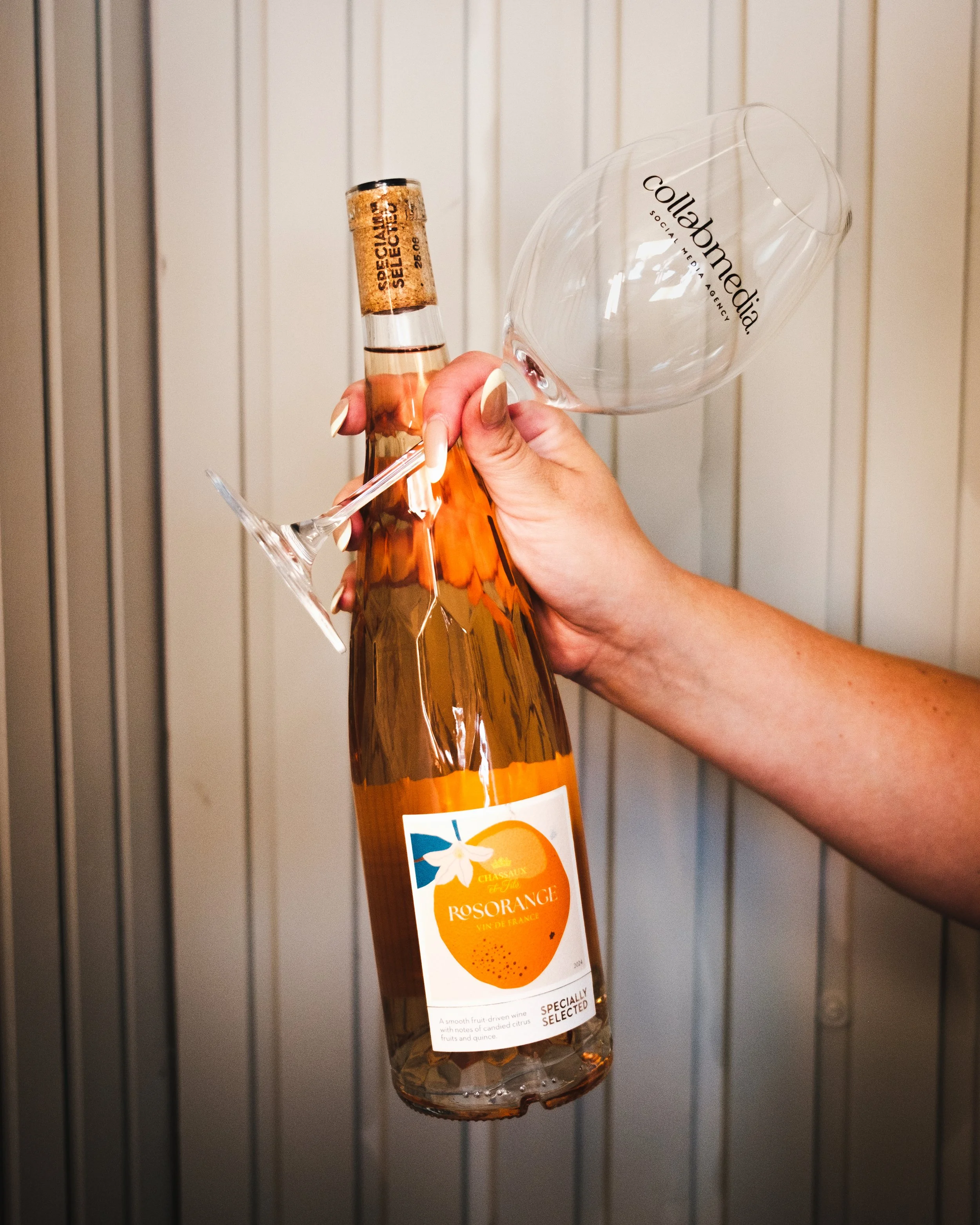 A hand holding a bottle of rosé wine with a wine glass balanced upside down on the neck of the bottle, against a vertical paneled background.