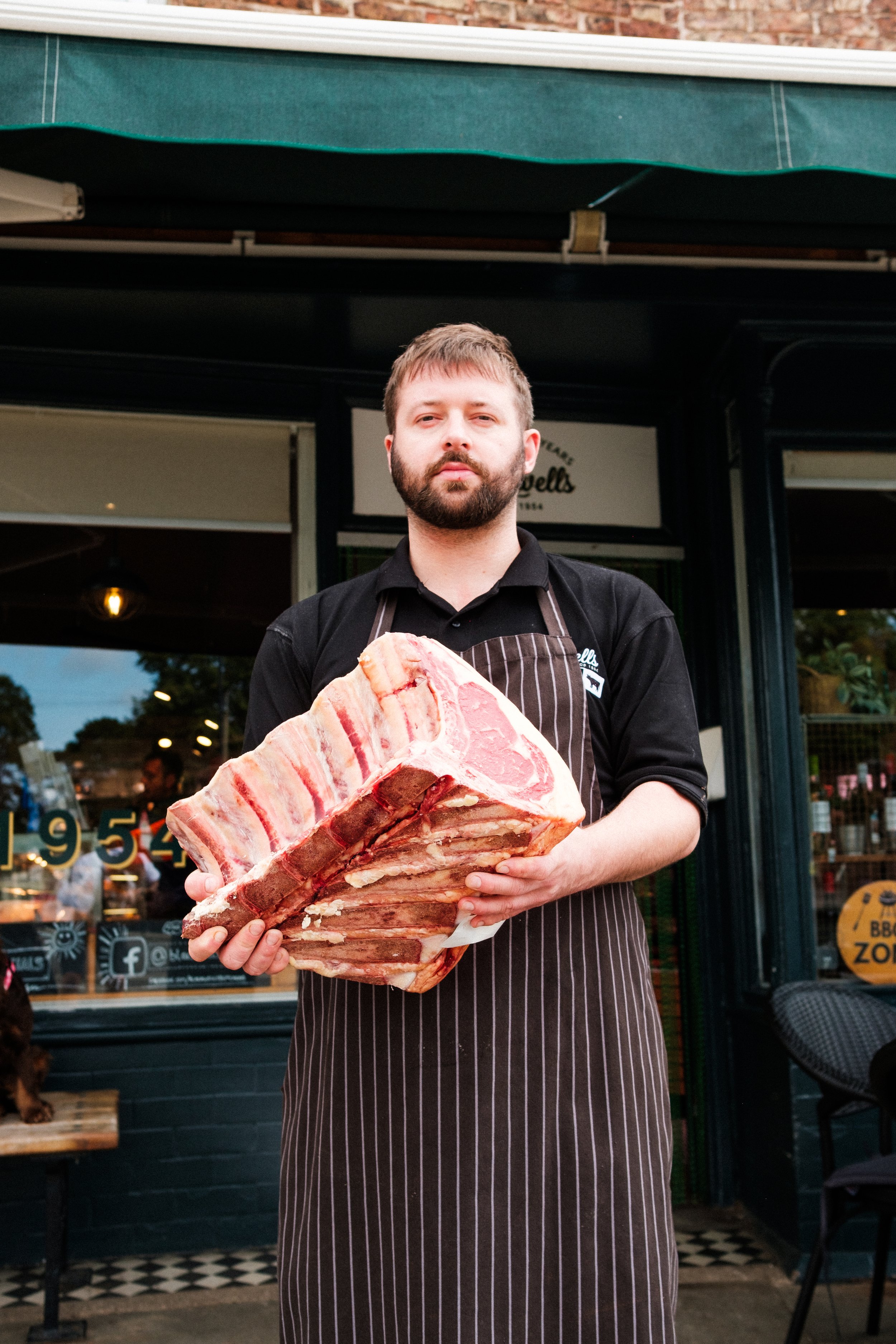 Man wearing an apron holding a large slab of raw bacon outside a storefront.