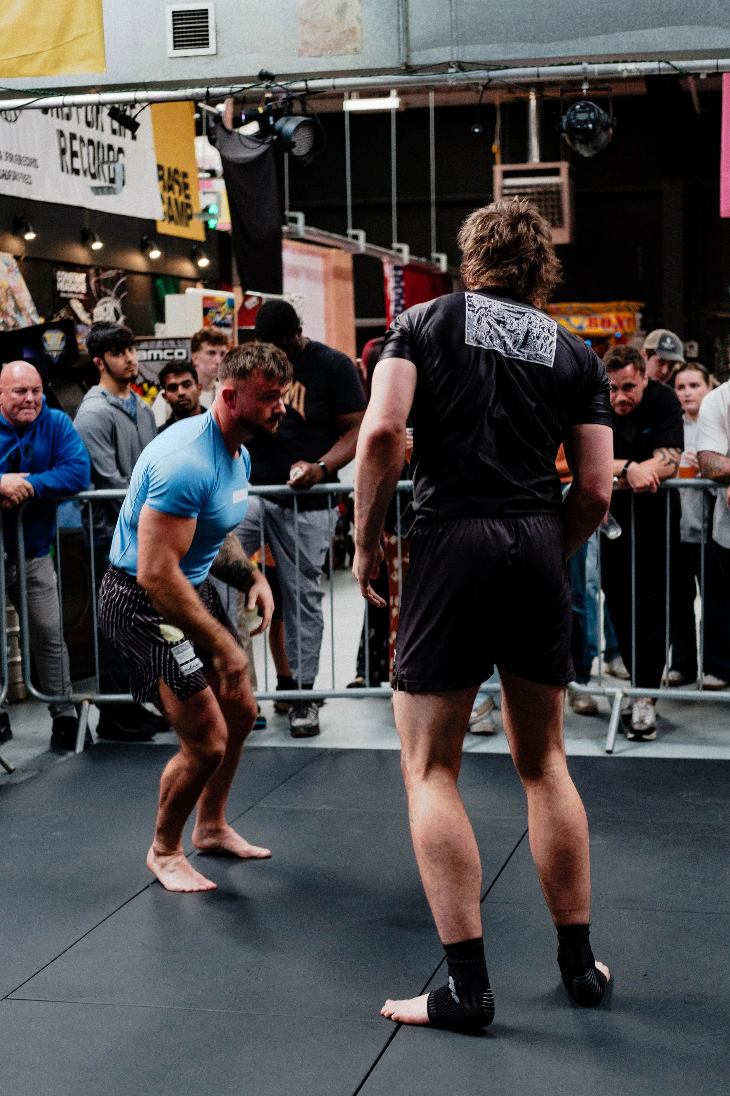 Two men barefoot on a black mat in a gym, engaged in a grappling or wrestling match, with an audience watching from behind a barrier.