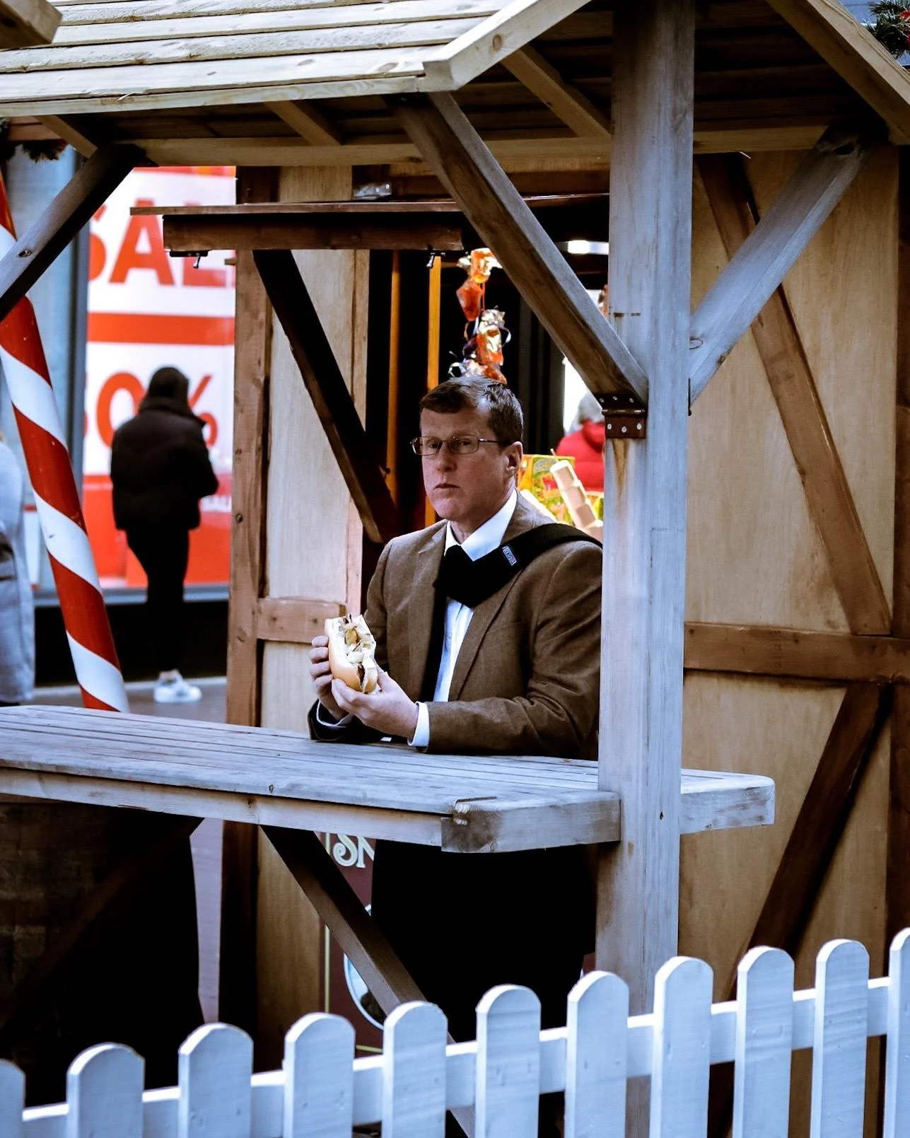 A man in a brown blazer and white shirt holding a hot dog inside a wooden food stand at an outdoor market.