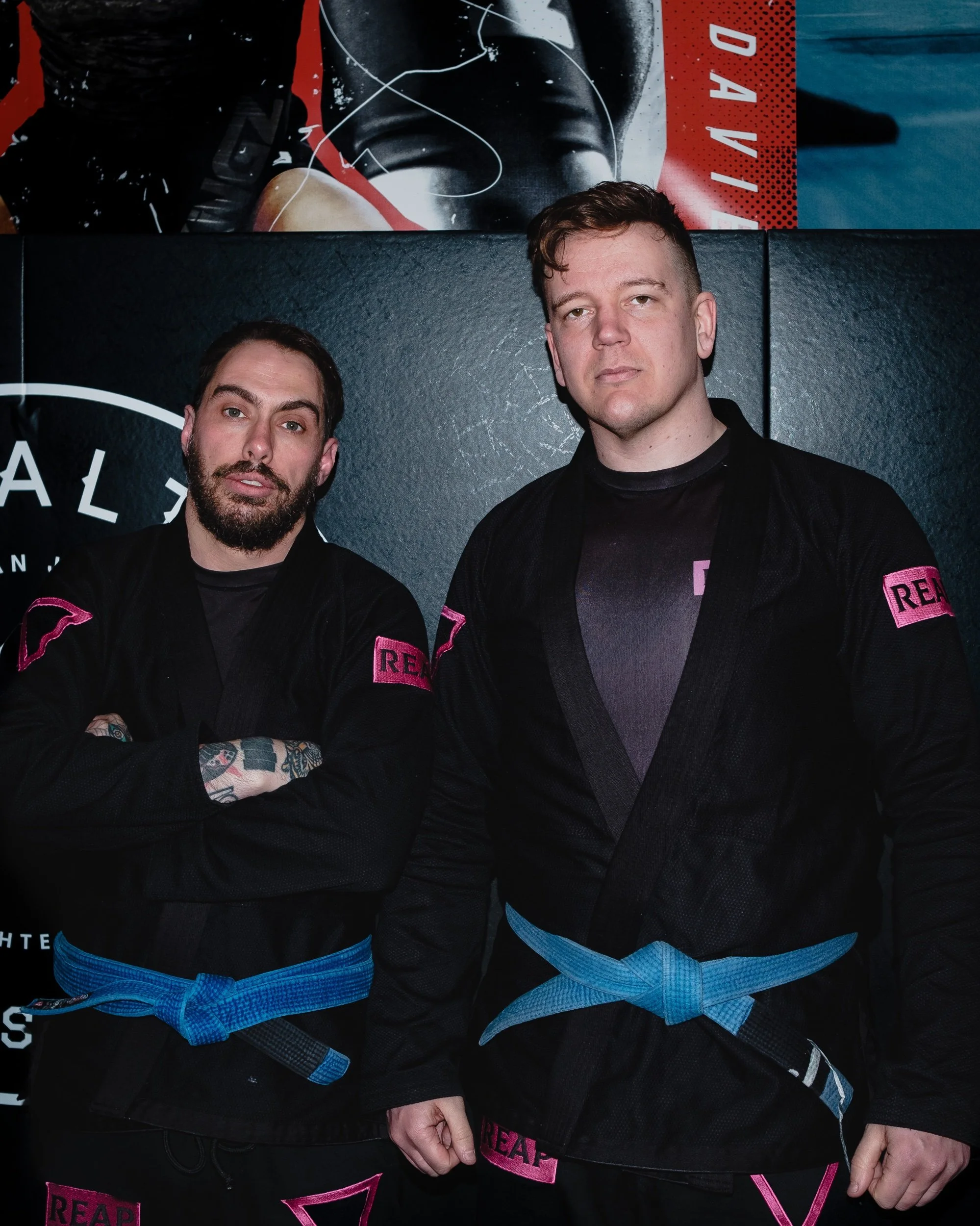 Two men stand side by side in martial arts uniforms with blue belts, black gis with pink patches. The man on the left has tattoos on his arms and a beard, while the man on the right has short hair and no beard. They are in a dojo or martial arts trai