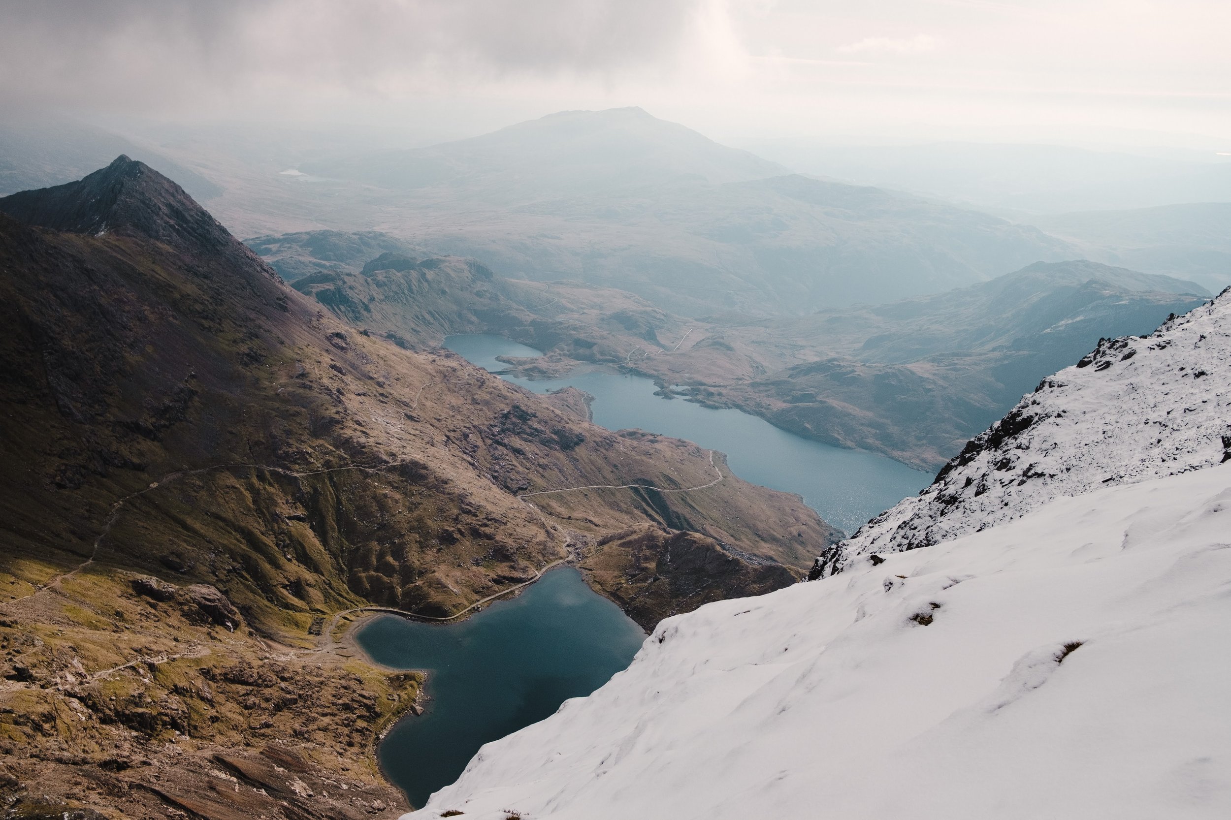 A mountainous landscape with snow-covered peaks in the foreground, rugged hills, and a series of lakes or reservoirs stretching into the distance under a cloudy sky.