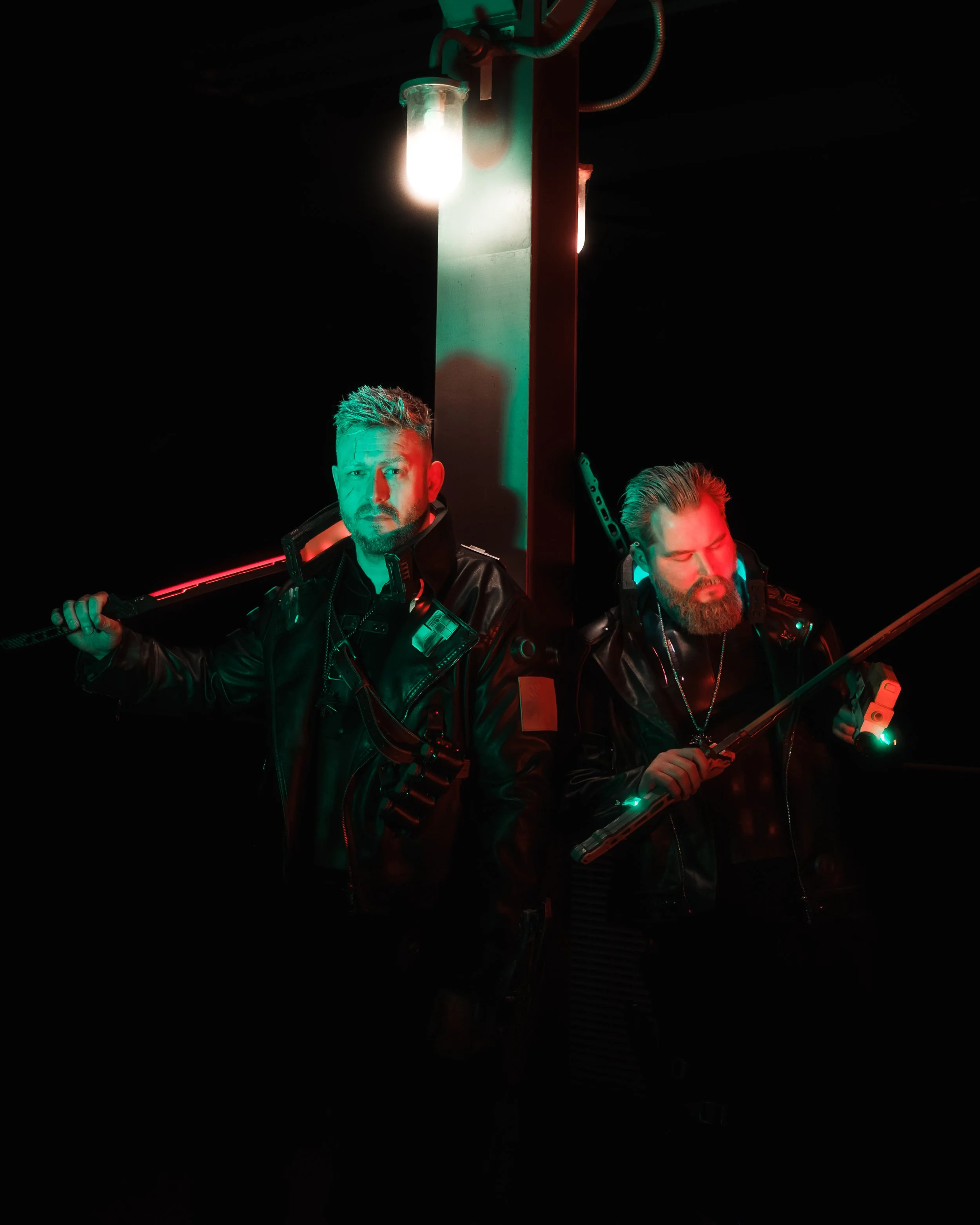 Two men wearing black leather jackets holding weapons, standing in a dark setting illuminated by green and red lights, with a bright light above them.