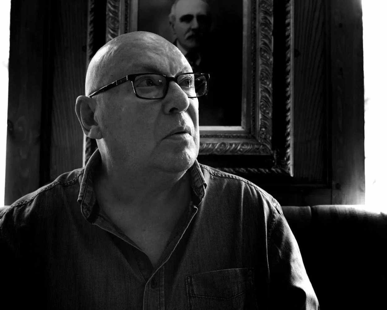 Black and white photo of a bald man wearing glasses, looking to the side, with a portrait of a person hanging behind him on a wooden-paneled wall.