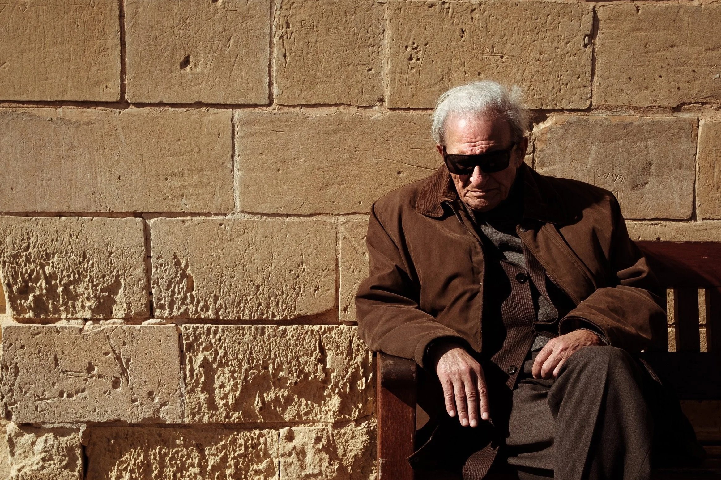 Older man with white hair wearing dark sunglasses and a brown jacket, sitting against a beige brick wall in sunlight.