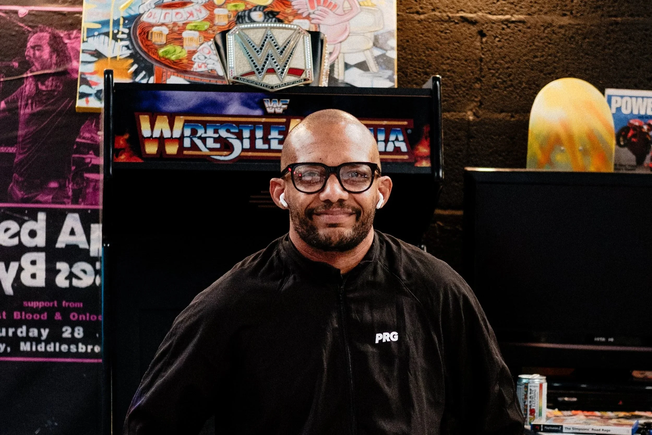 A man with glasses and earbuds standing in front of a WrestleMania arcade game, with a WWE championship belt on top of the game, and a colorful cartoon mural of Homer Simpson, Snoopy, and a pink creature playing poker behind him.
