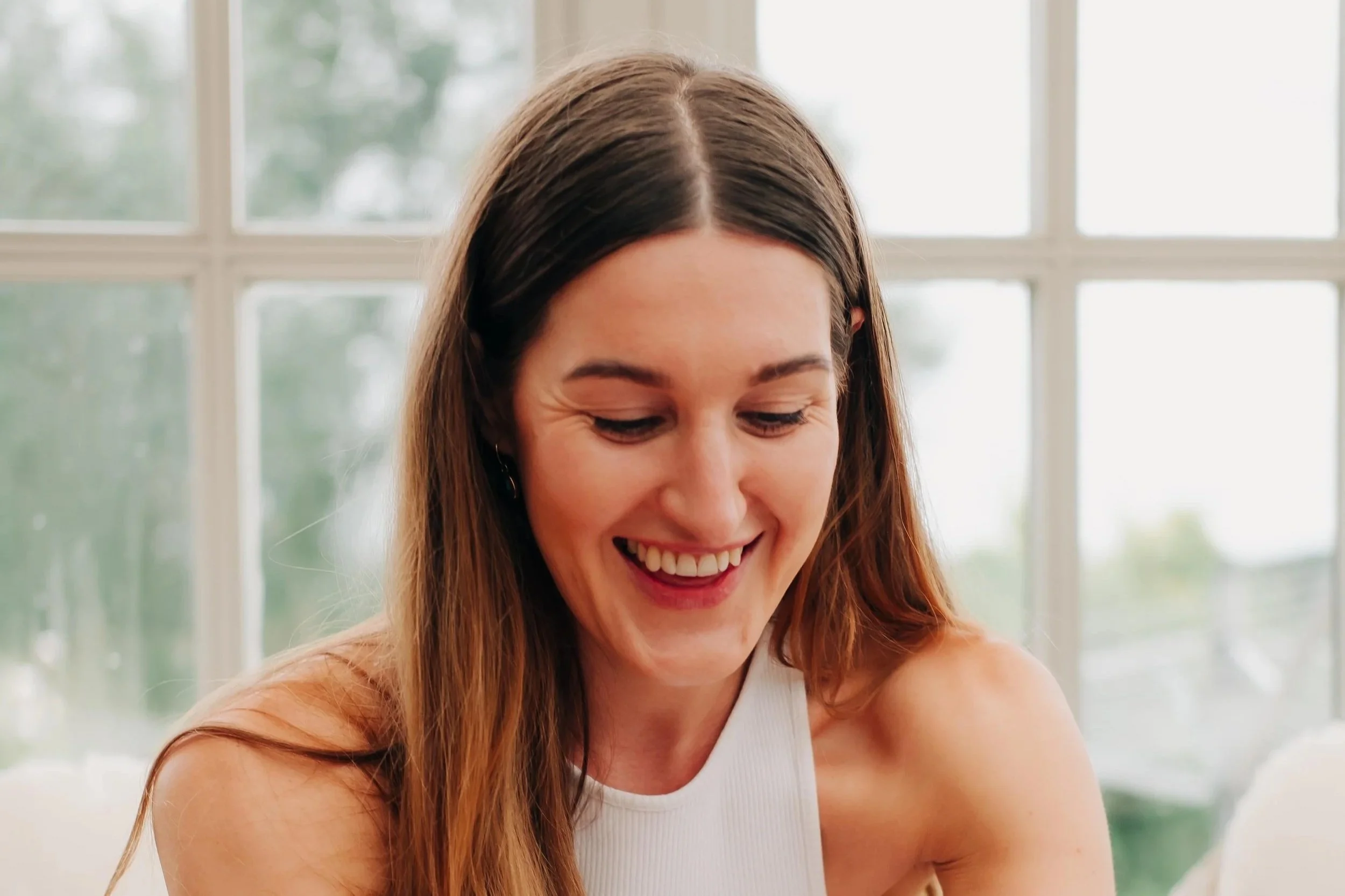 A woman with long brown hair, wearing a white sleeveless top and white skirt, is opening a bottle of champagne in a bright room with large windows and a blurred outdoor view.