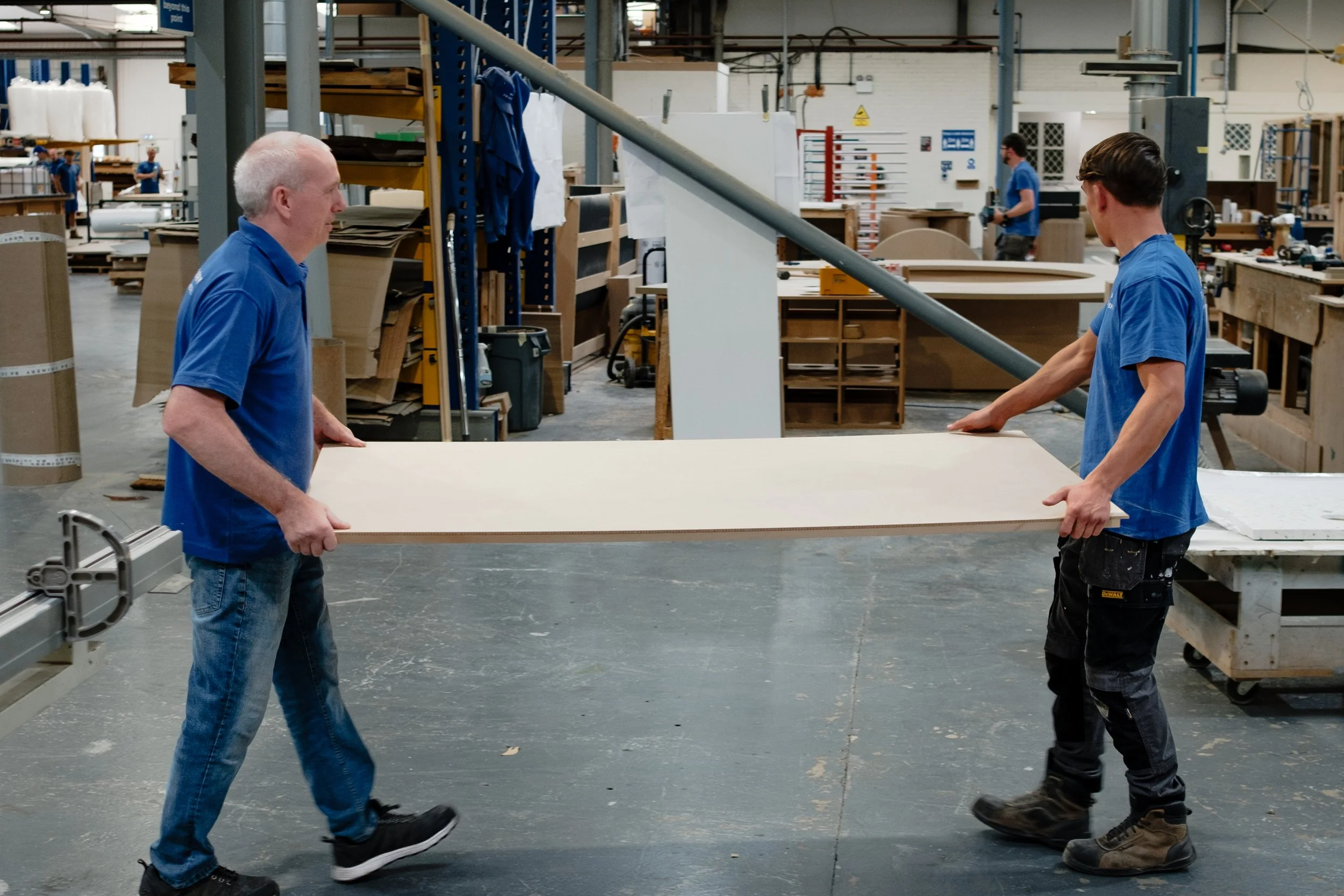 Two workers in blue shirts are passing a large sheet of wood in a woodworking shop. The shop has various tools, shelves, and other workers in the background.